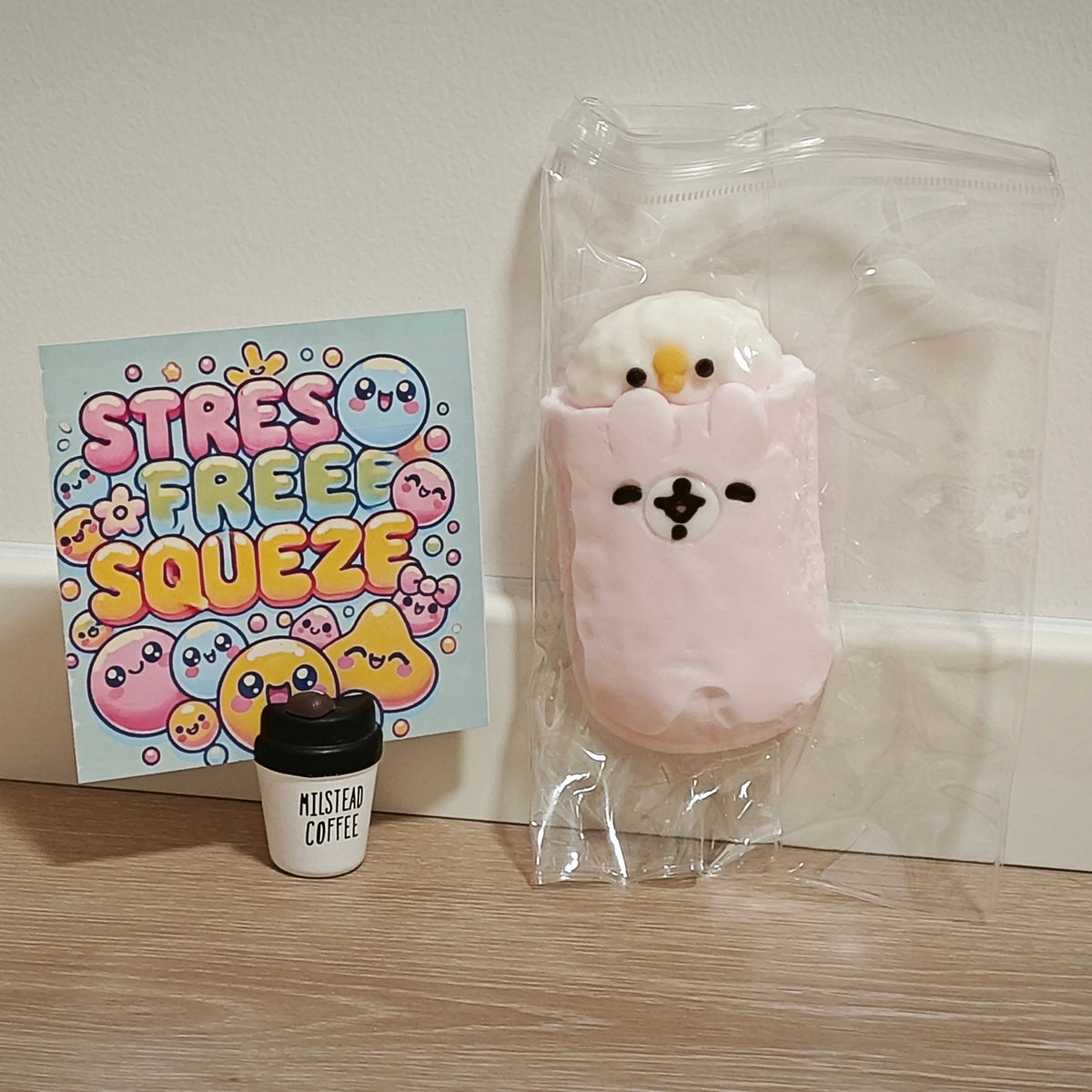 A pink plush stress toy shaped like a bird peeking out of a coffee cup, sealed in a clear plastic bag next to a colorful 'STRESS FREE SQUEEZE' card and a miniature coffee cup accessory on a wooden surface.
