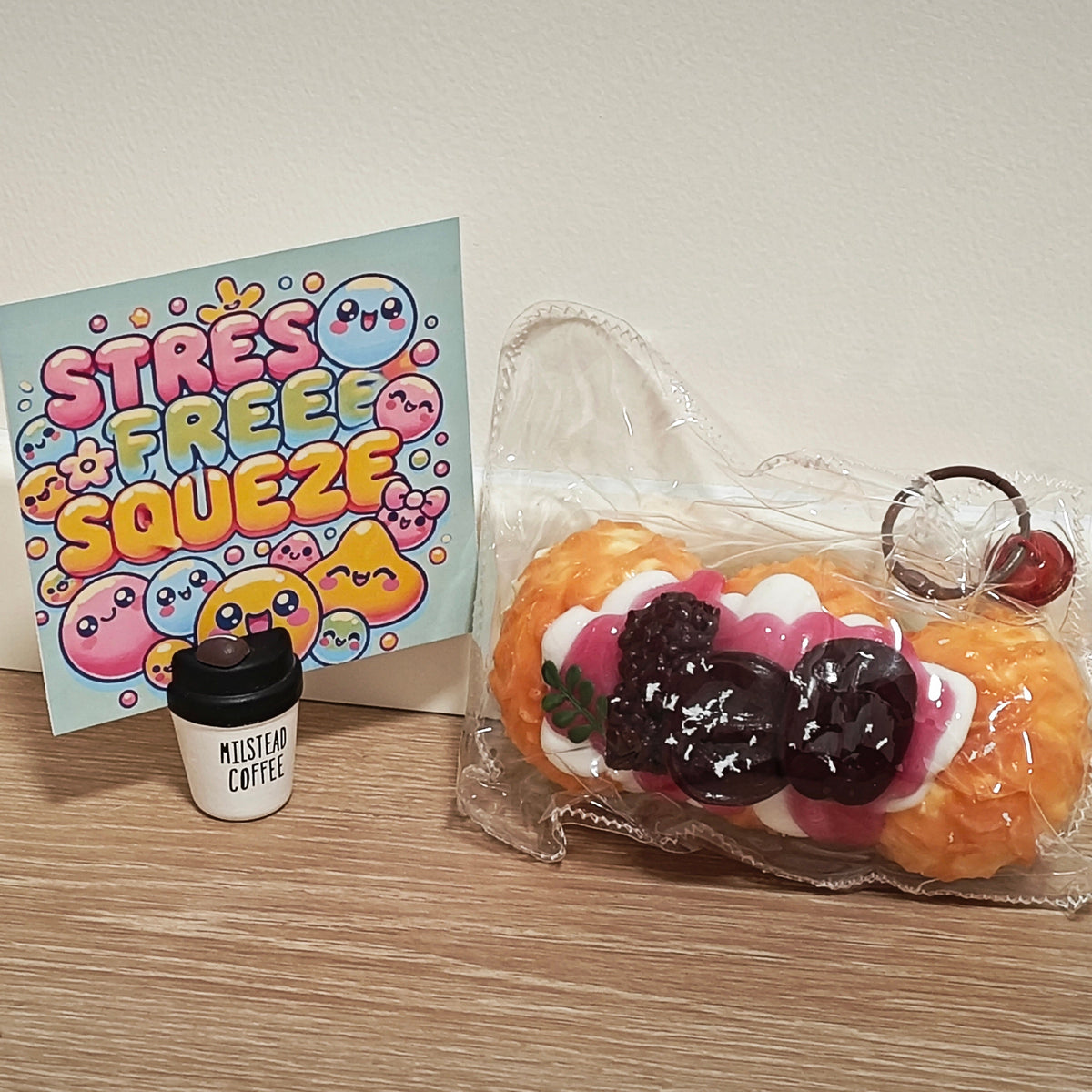 A novelty squishy toy shaped like a strawberry pastry in a clear plastic bag, displayed on a wooden surface next to a small cup labeled 'Mister Coffee' and a card that reads 'Stress Free Squeeze'.