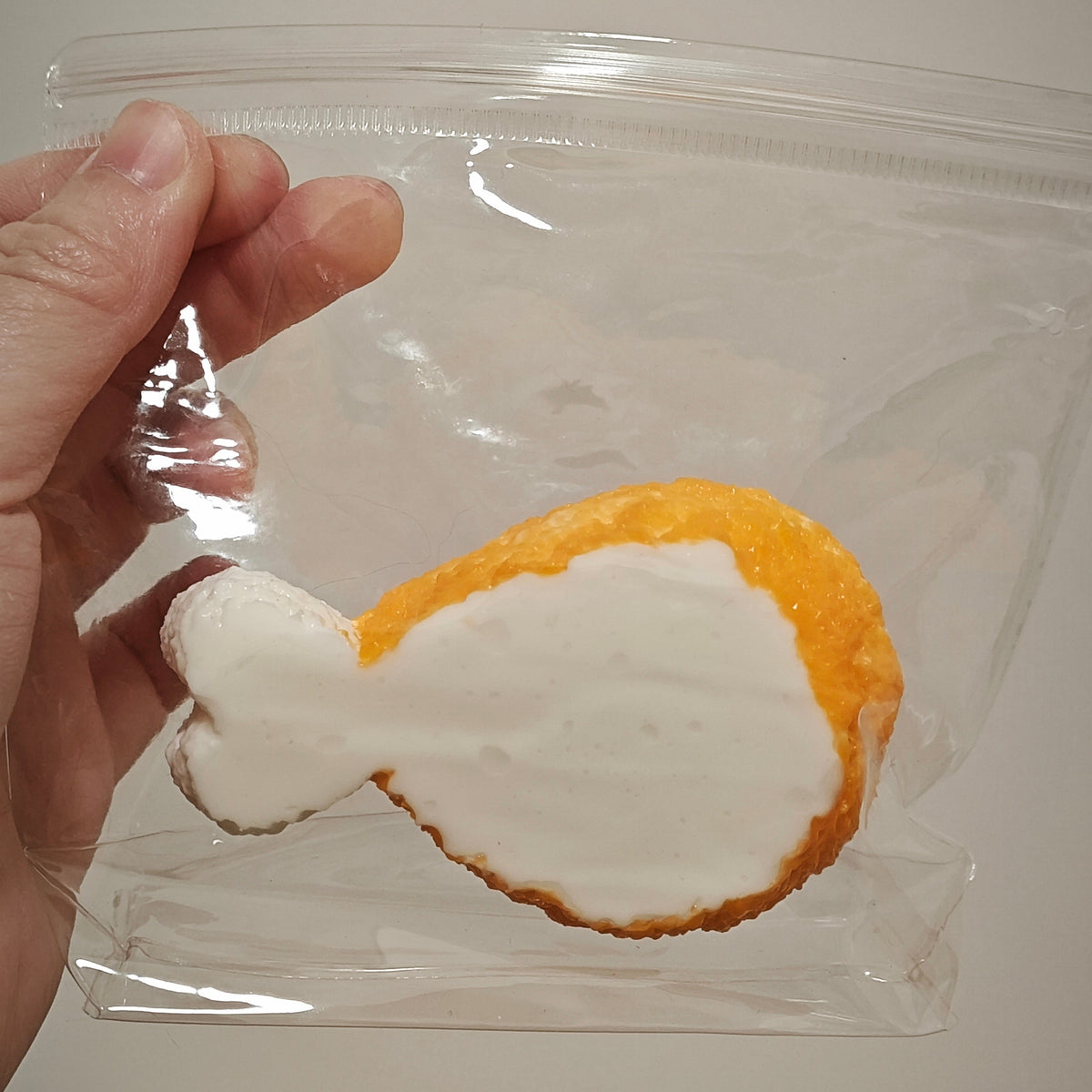 A hand holding a clear plastic zip-top bag containing a novelty fried chicken leg shaped cookie or candy with white frosting-like filling, against a plain light background.