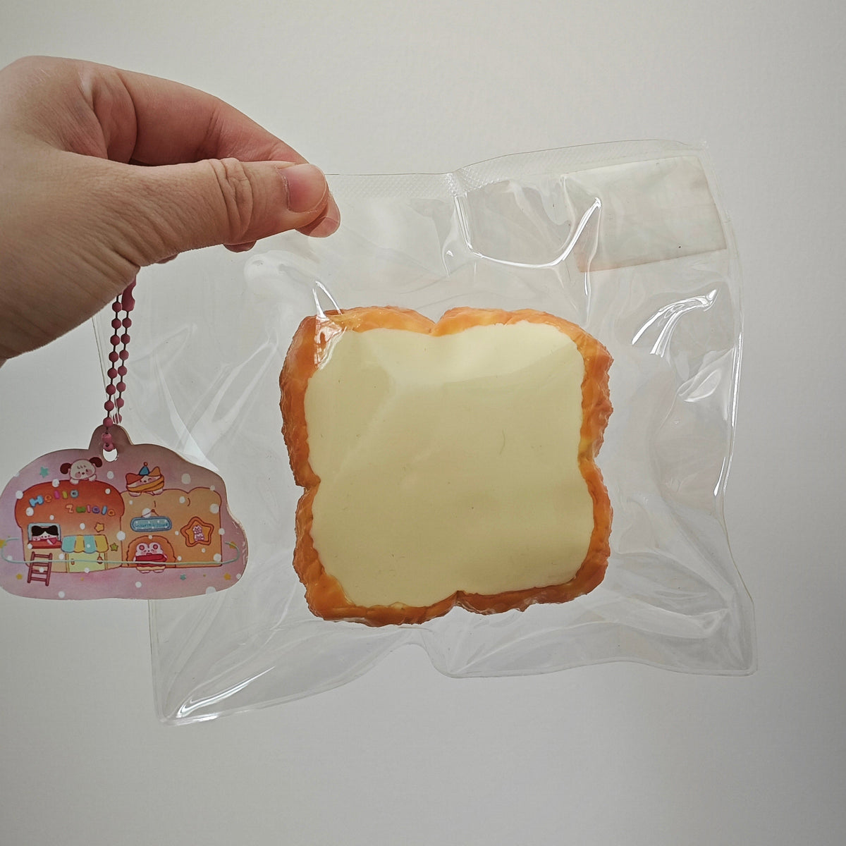 A hand holds a clear plastic bag containing a bread-slice-shaped item with a pale yellow center and golden-brown edges, next to a small pink illustrated keychain charm.