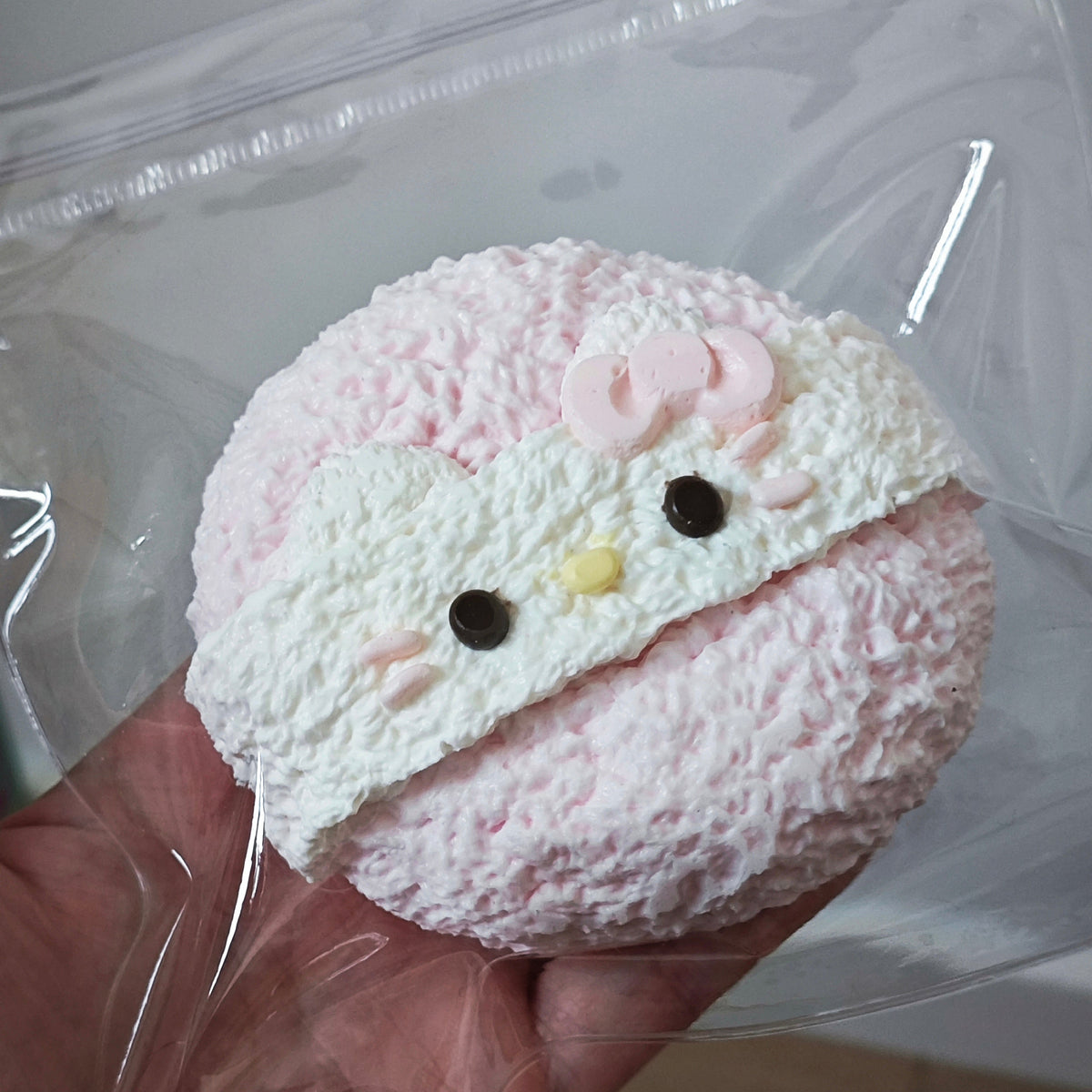 A pink and white textured Hello Kitty-themed squishy toy held in a hand inside a clear plastic bag.