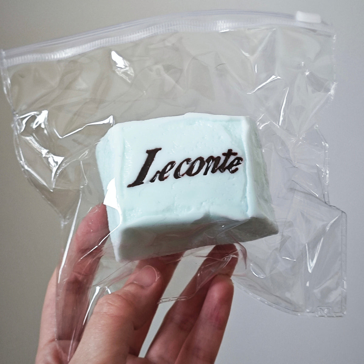 A hand holding a clear plastic zip-top bag containing a pale mint green rectangular product with the brand name 'Leconte' printed in dark brown script on the front, against a plain light gray background.