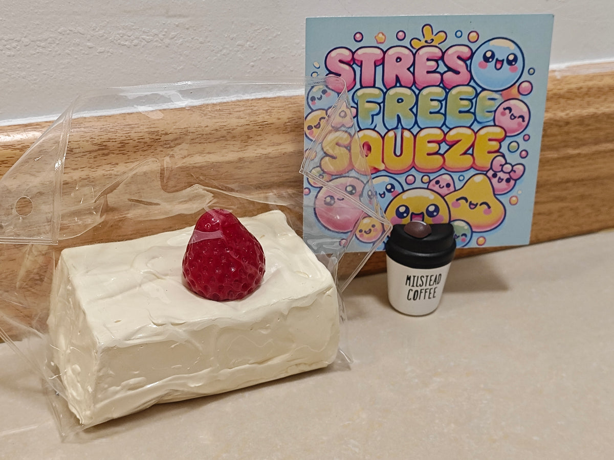 A miniature toy set featuring a cream-colored cake with a red strawberry, a small coffee cup labeled 'Mistaken Coffee,' and a colorful card that reads 'STRESS FREE SQUEEZE' with cartoon characters.