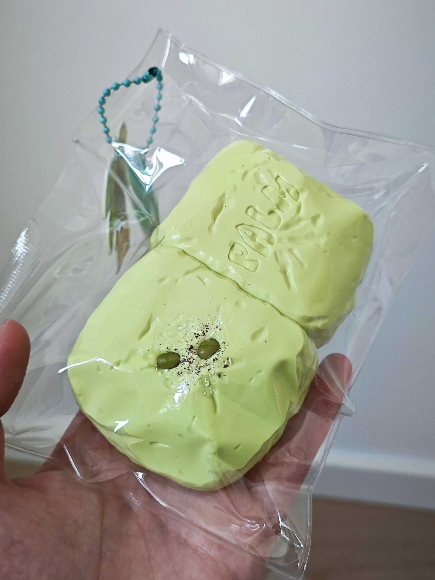 A hand holding a clear plastic bag containing a pale yellow-green novelty soap or bath bomb shaped like a sliced fruit, with a small green bead keychain attached.