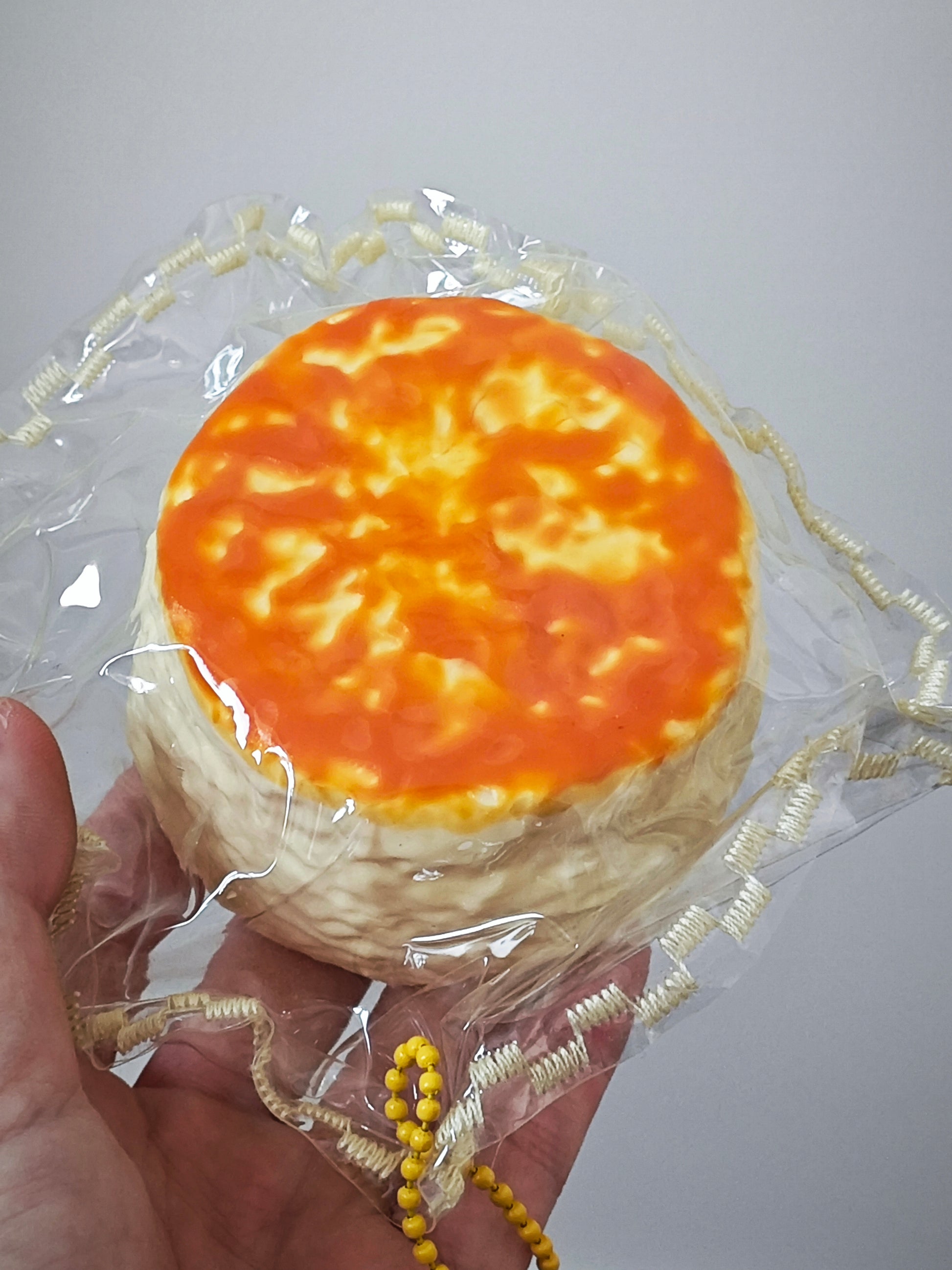 A hand holding a small round novelty item shaped like a baked cake or pastry, wrapped in clear plastic with a decorative beige ribbon and yellow bead string, against a plain light gray background.