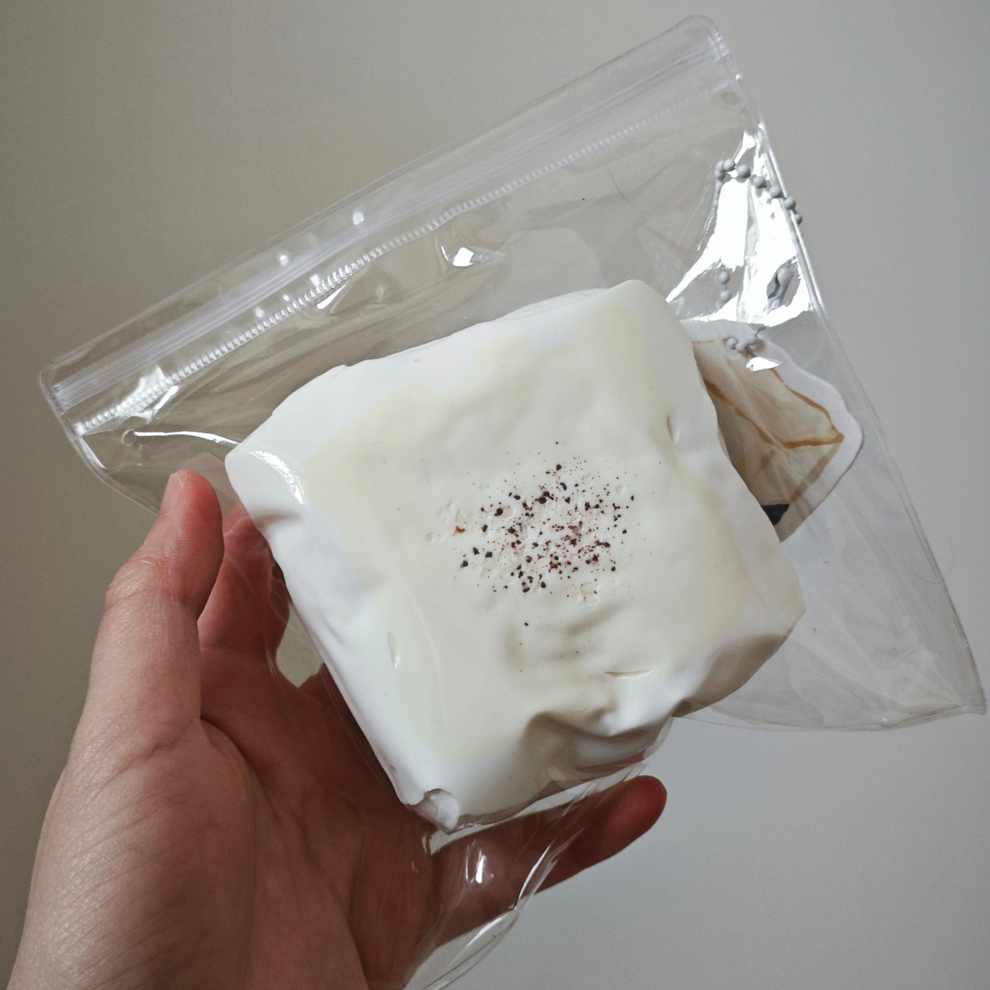 A hand holding a clear plastic zip-top bag containing a white, square, crumpled item with a small sprinkle of dark seasoning on top, against a plain light gray background.