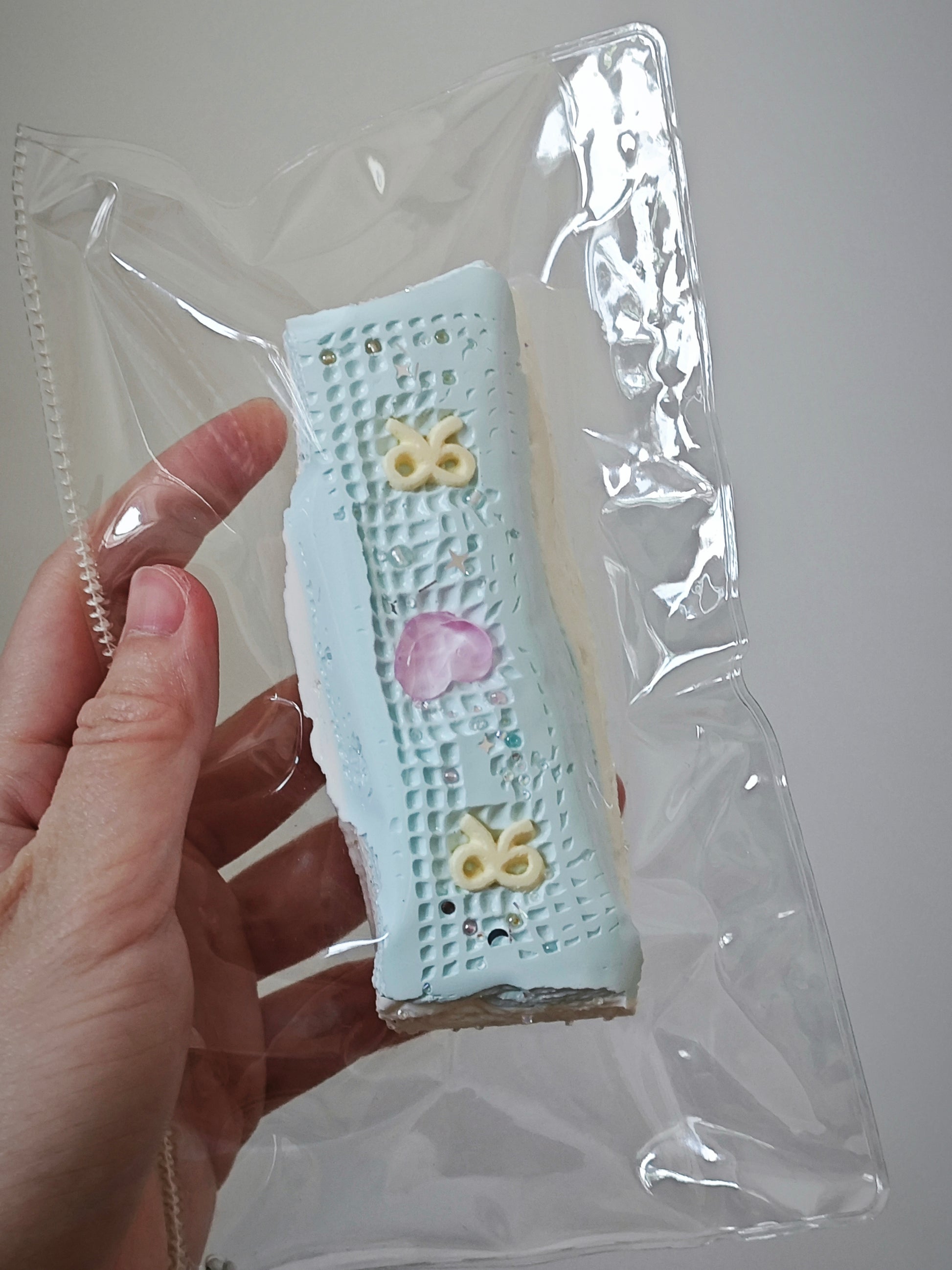 A hand holding a small rectangular pastel item inside a clear plastic bag. The item appears light blue with a grid texture and decorative pastel motifs, including yellow and pink accents.