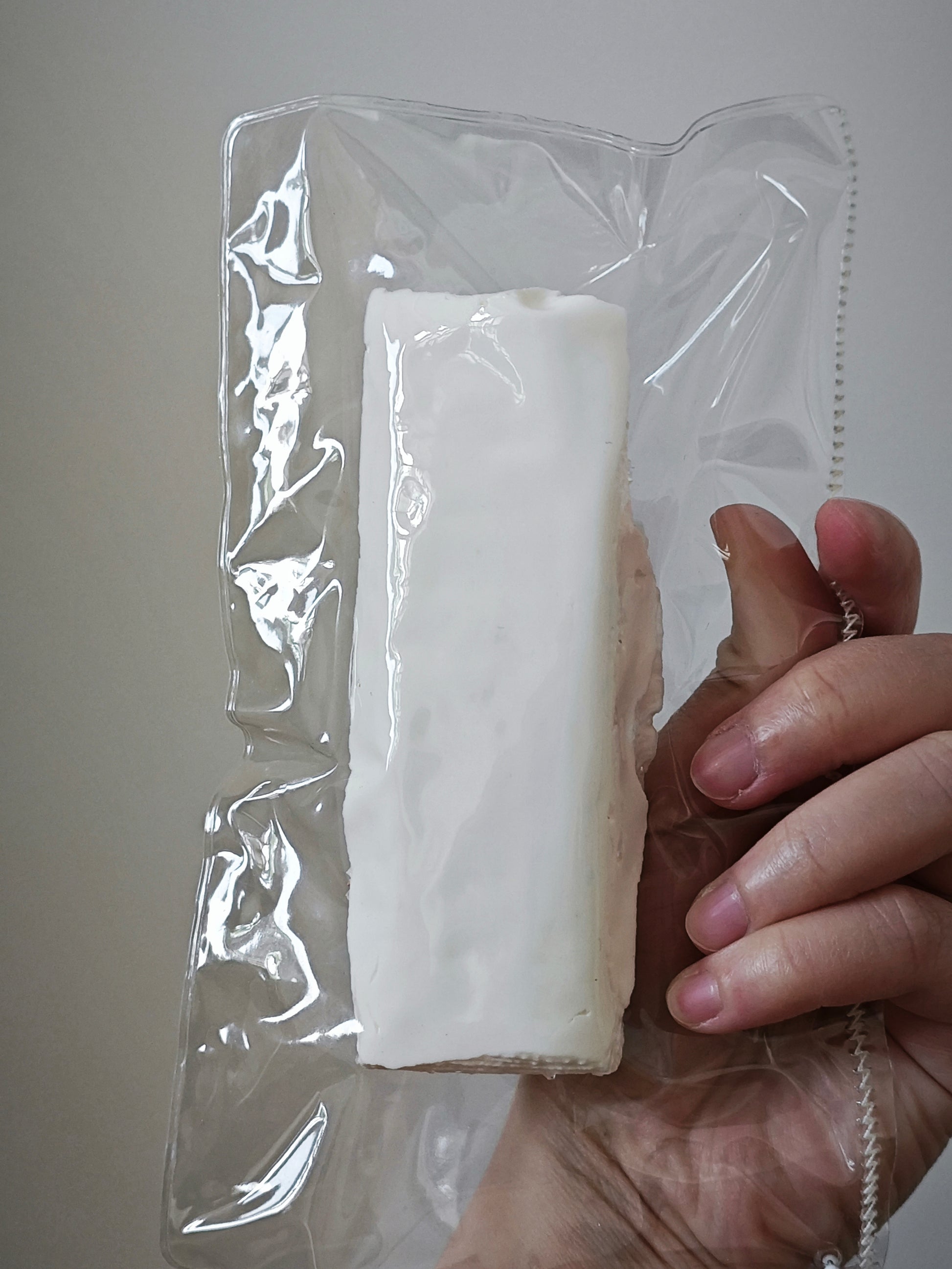 A hand holding a vacuum-sealed clear plastic bag containing a white rectangular block, photographed against a plain light gray background.