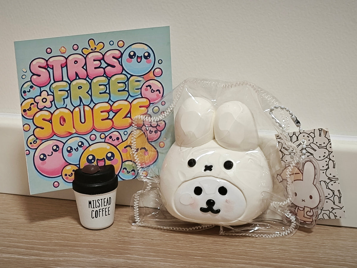 A white ceramic rabbit-shaped stress toy in clear plastic packaging, displayed next to a colorful 'STRESS FREE SQUEEZE' card and a small 'MISTBEAD COFFEE' cup on a wooden surface against a beige wall.