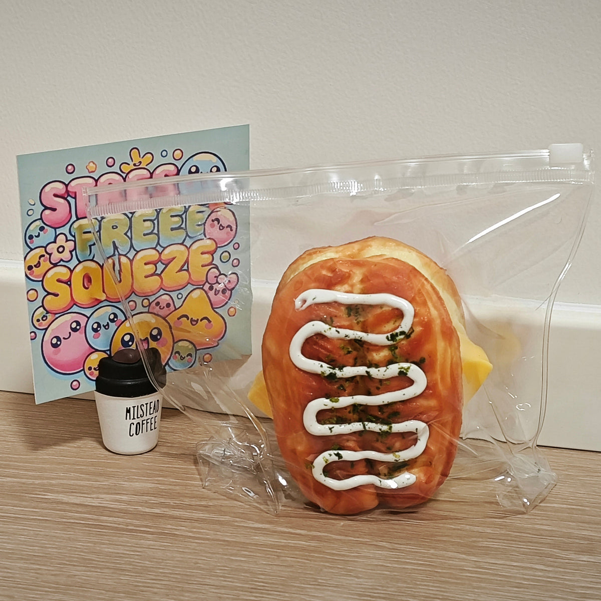 A novelty squishy toy shaped like a sausage on a bun, sealed in a clear plastic bag, displayed on a wooden surface next to a small coffee cup prop and a colorful 'Stress Free Squeezie' card.