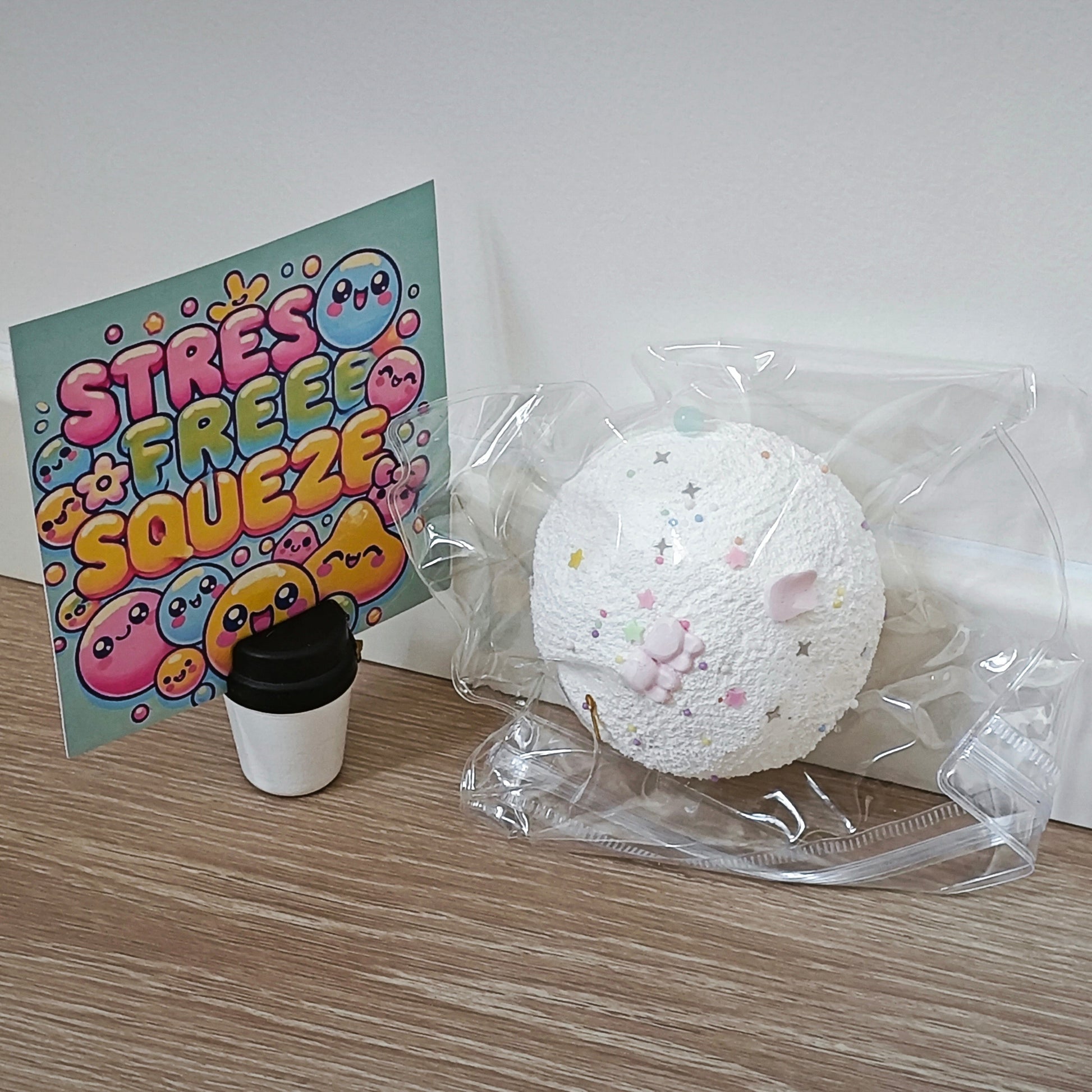 A white textured squishy toy with pastel pink and yellow details sits in a clear plastic bag on a light wood floor against a white wall. A colorful card reading 'STRESS FREE SQUEEZE' stands on a small black-and-white cup next to the packaged toy.