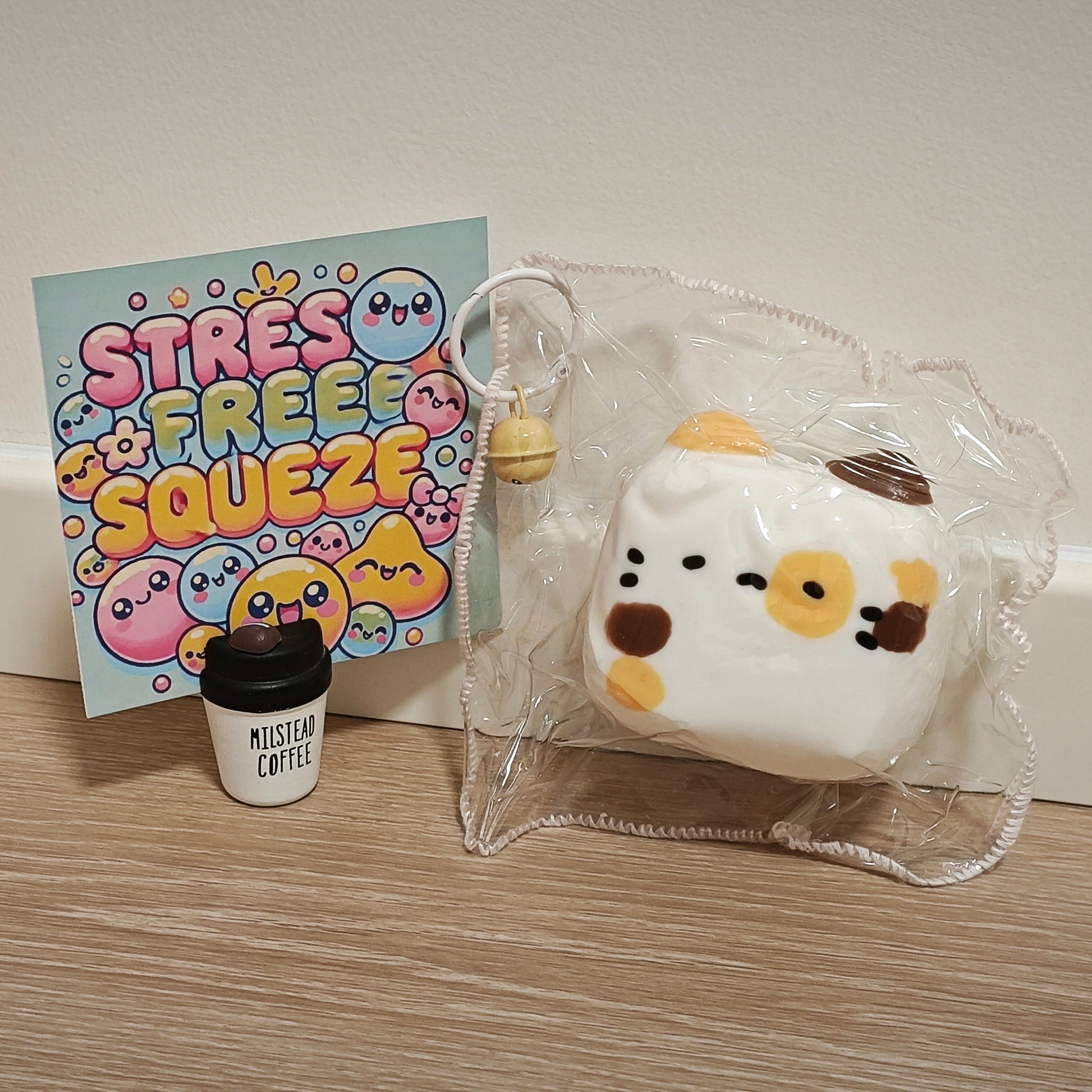 A white square squishy toy with a cow face design, sealed in a clear plastic bag, displayed next to a colorful 'STRESS FREE SQUEEZE' card and a miniature coffee cup on a wooden surface against a plain wall.