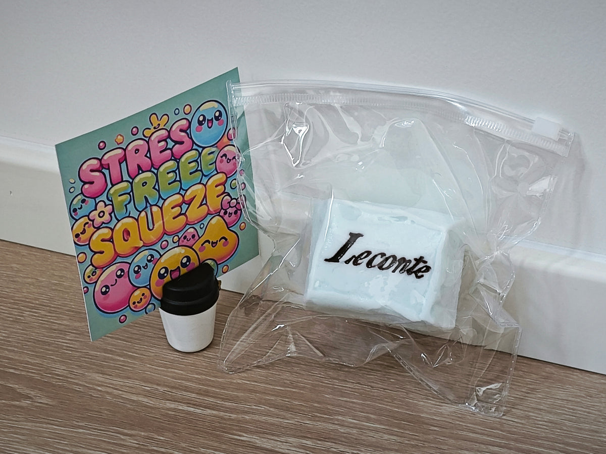 A white rectangular item labeled 'Leconte' inside a clear plastic bag, next to a colorful card that reads 'STRESS FREE SQUEEZF' with cartoon faces, and a small black-and-white cup, on a wood surface against a white wall.