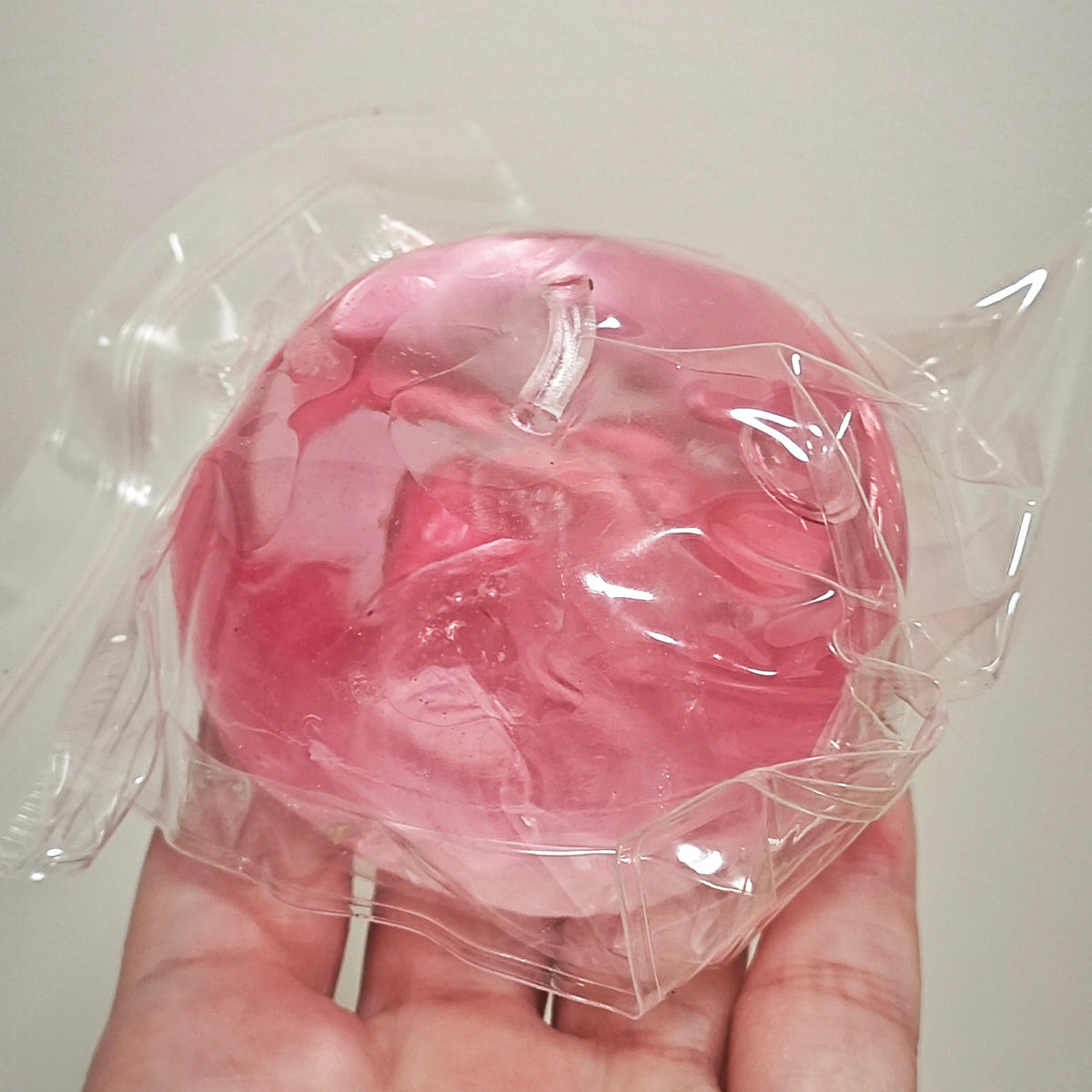 A hand holding a pink, round, translucent gel-like product sealed in clear plastic packaging against a plain light background.