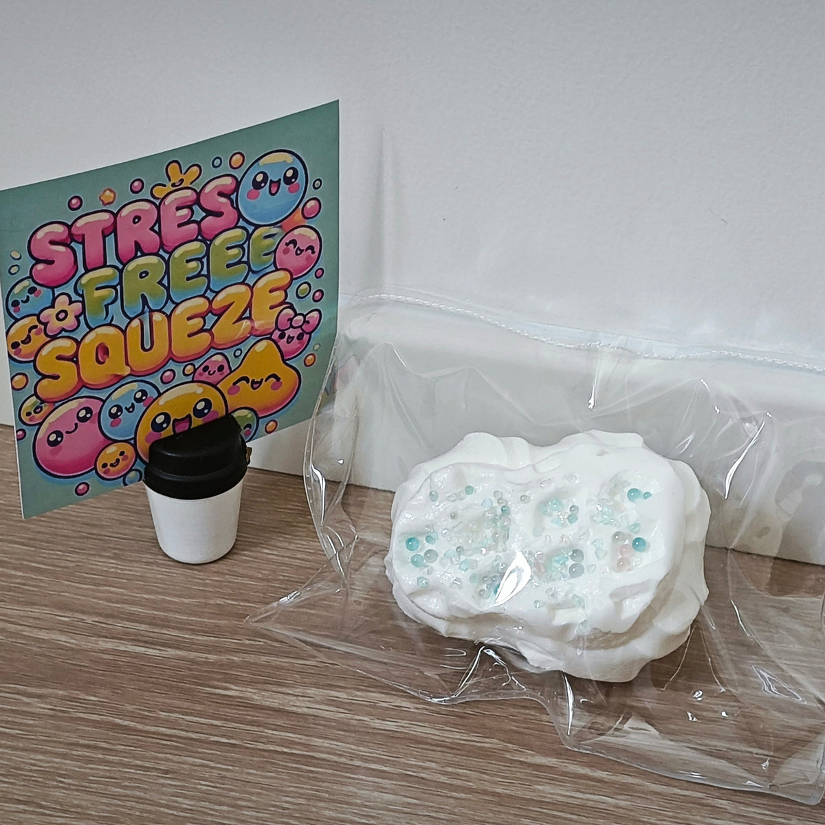A white, textured stress toy with embedded blue glitter sits in a clear plastic bag on a wooden floor, next to a small black-and-white coffee cup and a colorful card that reads 'STRESS FREE SQUEEZEE'.