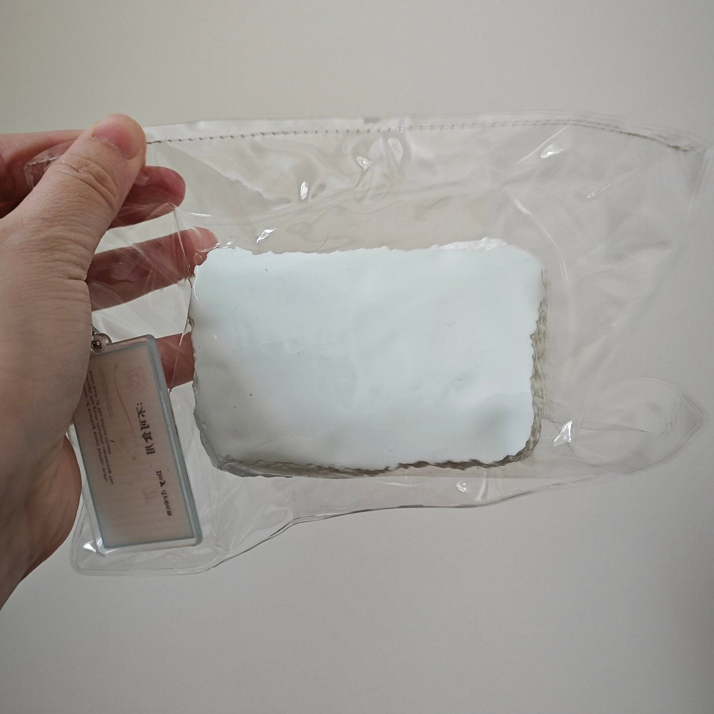 A hand holding a clear plastic zip pouch containing a white rectangular item, with a small hanging tag attached, against a plain light background.