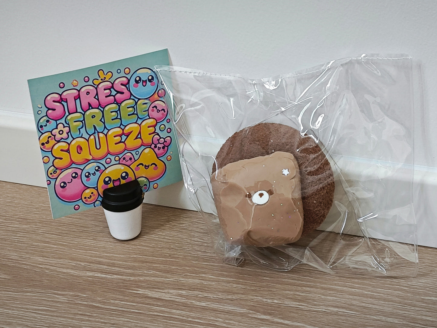 A squishy toy shaped like a slice of toast or pastry with a brown cookie-like base and beige frosting, sealed in a clear plastic bag. A colorful card reading 'STRESS FREE SQUEEZA' and a small takeaway coffee cup accessory sit beside it on a light wood surface against a white wall.