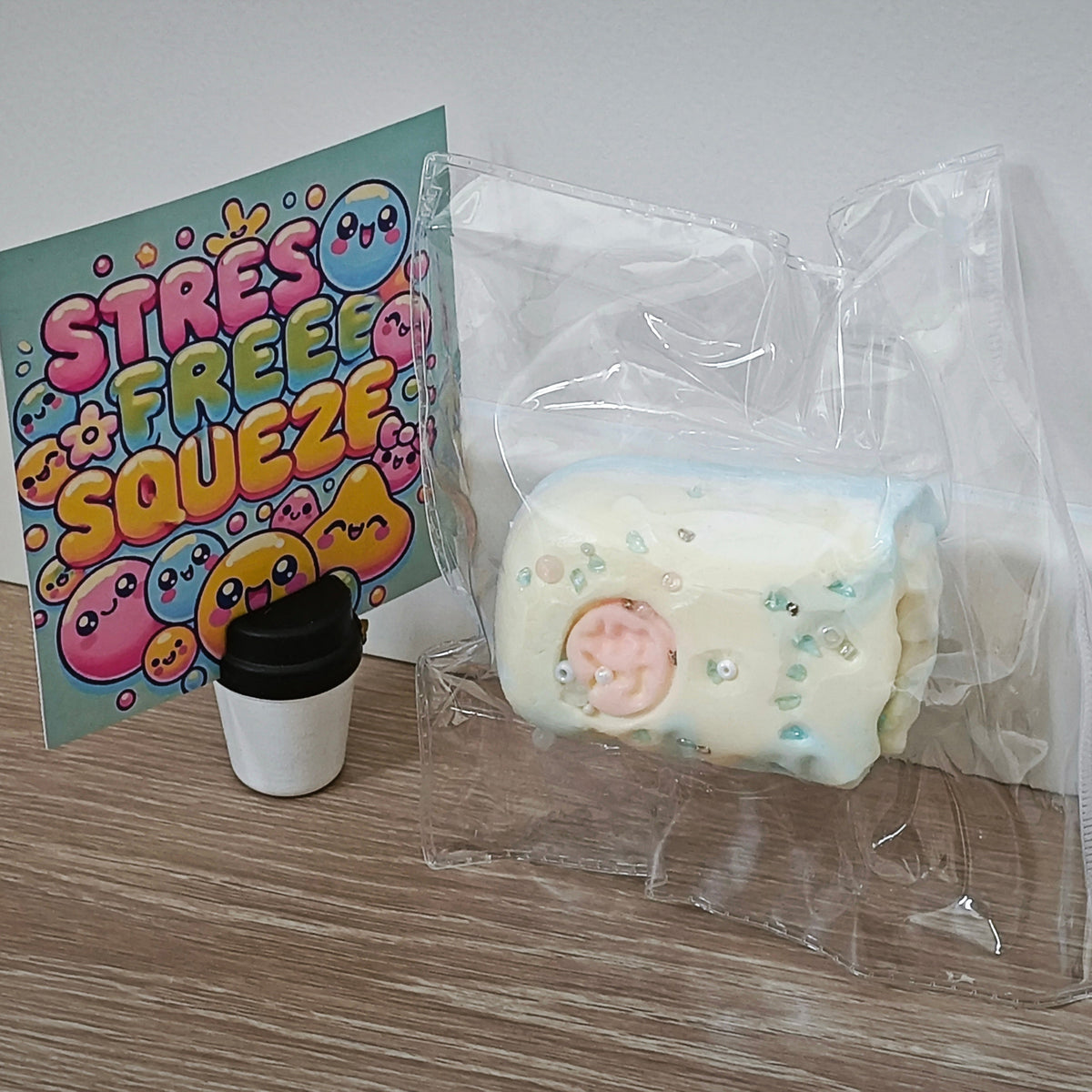 A packaged squishy toy sits on a wood floor against a white wall, next to a colorful 'STRESS FREE SQUEEZE' card and a small black-and-white cup-shaped accessory. The main product is wrapped in a clear plastic bag and appears to be a cream-colored cylindrical squishy with pastel blue and pink details.
