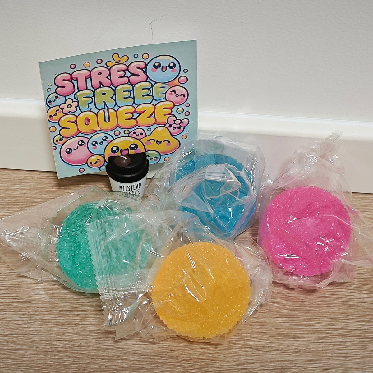 Four round, textured stress balls in teal, blue, yellow, and pink, each individually wrapped in clear plastic, arranged on a light wood surface in front of a colorful 'STRESS FREE SQUEZE' card and a small black jar.