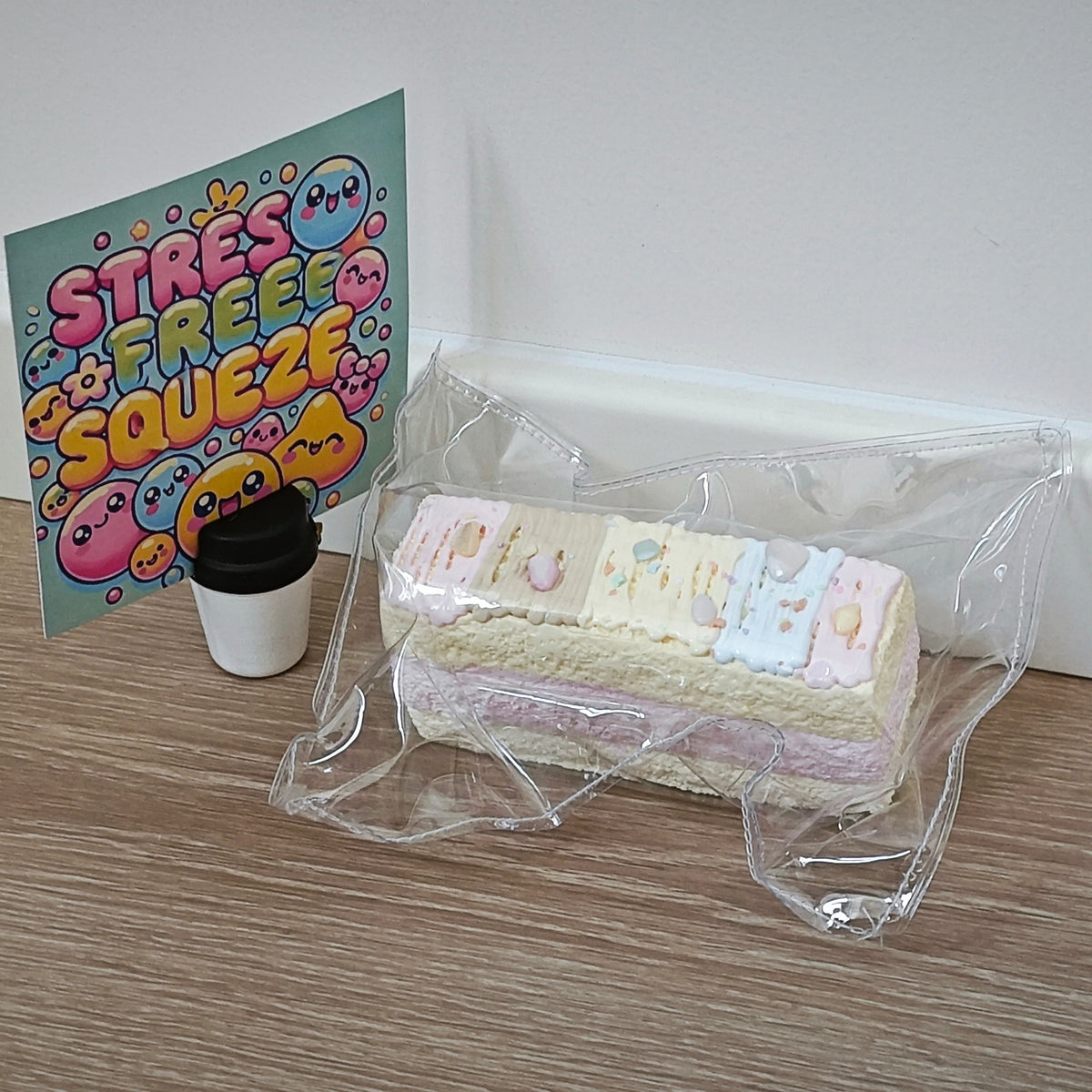 A pastel-colored layered cake squishy toy in a clear plastic bag sits on a wooden floor next to a colorful 'Stress Free Squeeze' card and a small coffee cup accessory.