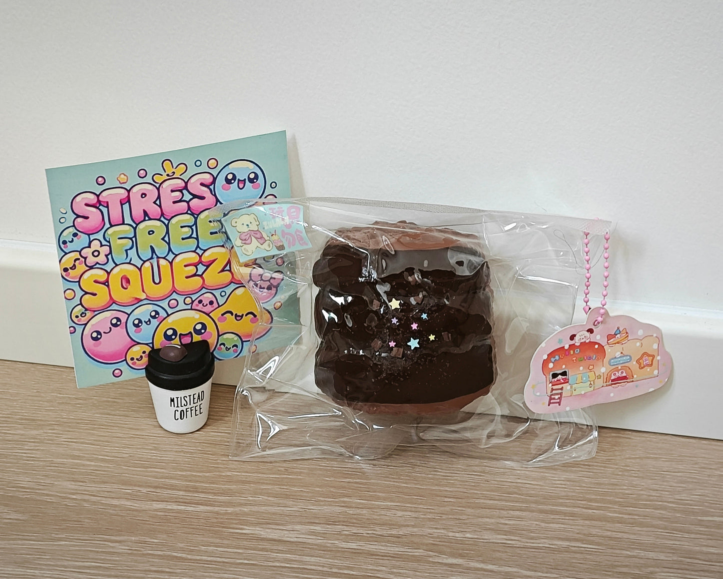 A chocolate cake-shaped squishy toy in clear plastic packaging sits on a light wood surface against a white wall, alongside a colorful 'Stress Free Squeeze' card, a small 'Mistress Coffee' cup, and a pink cloud-shaped tag.