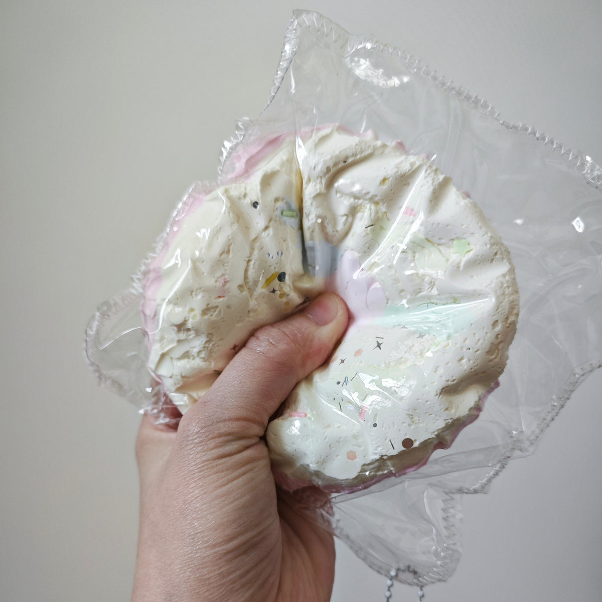 A hand holding a round, frosted donut-shaped squishy toy inside a clear plastic bag with a small character keychain attached, against a plain light gray background.