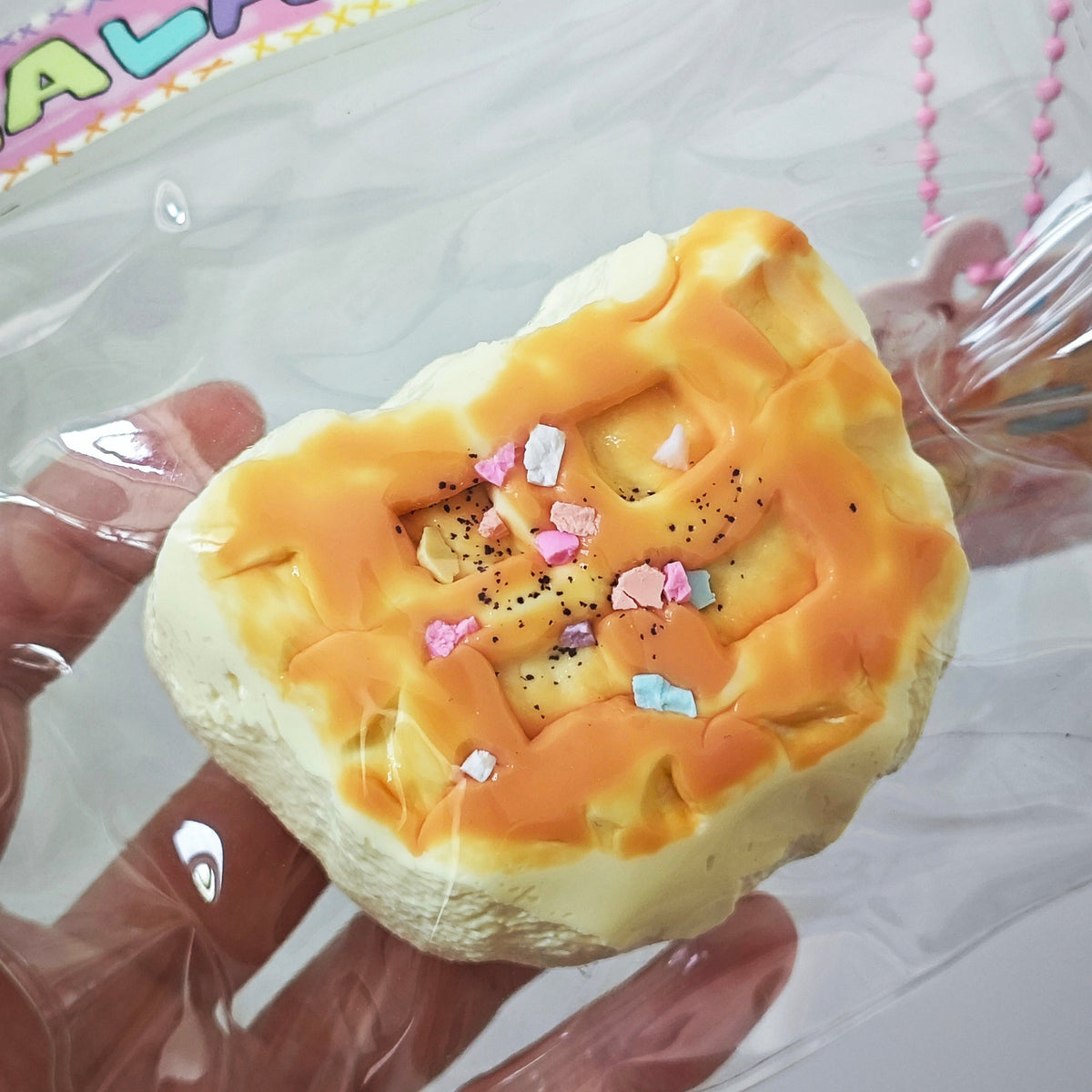 A hand wearing a clear plastic glove holds a baked pastry-shaped squishy toy with orange frosting, cream-colored sides, and small pink and blue sprinkles. The item is packaged in a clear plastic sleeve with a decorative header card.