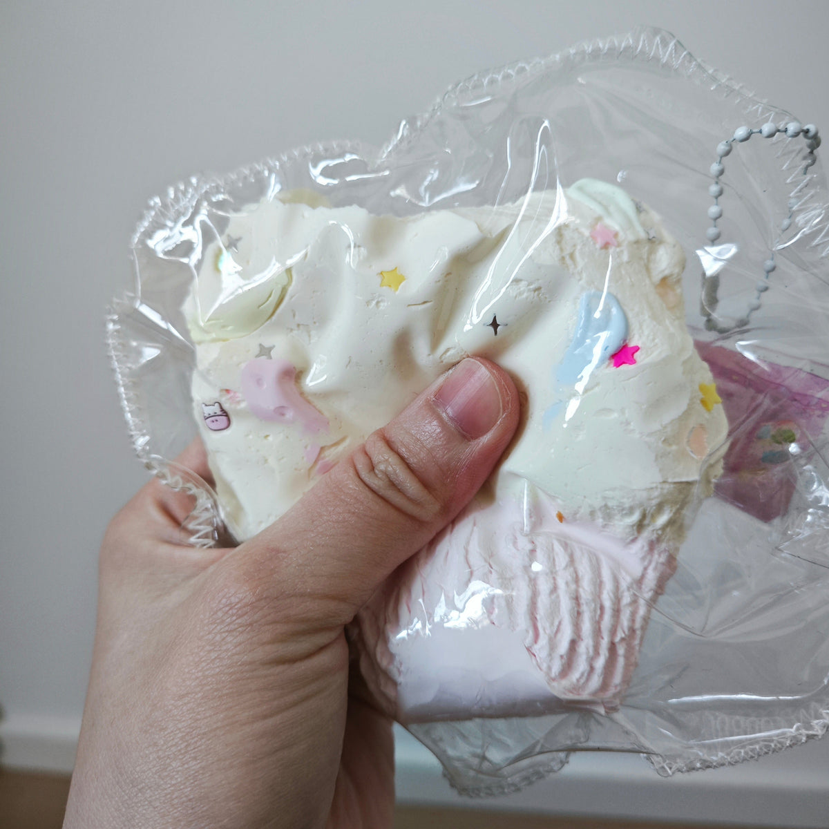 A hand holding a squishy toy inside a clear plastic bag. The squishy is primarily off-white with pastel pink and light blue accents, decorated with tiny star and unicorn motifs.