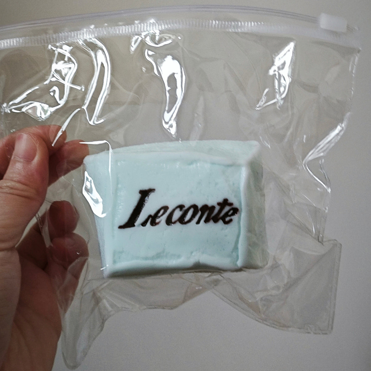 A hand holding a clear plastic zip bag containing a pale blue rectangular item with the word 'Leconte' printed in black script, against a plain light background.