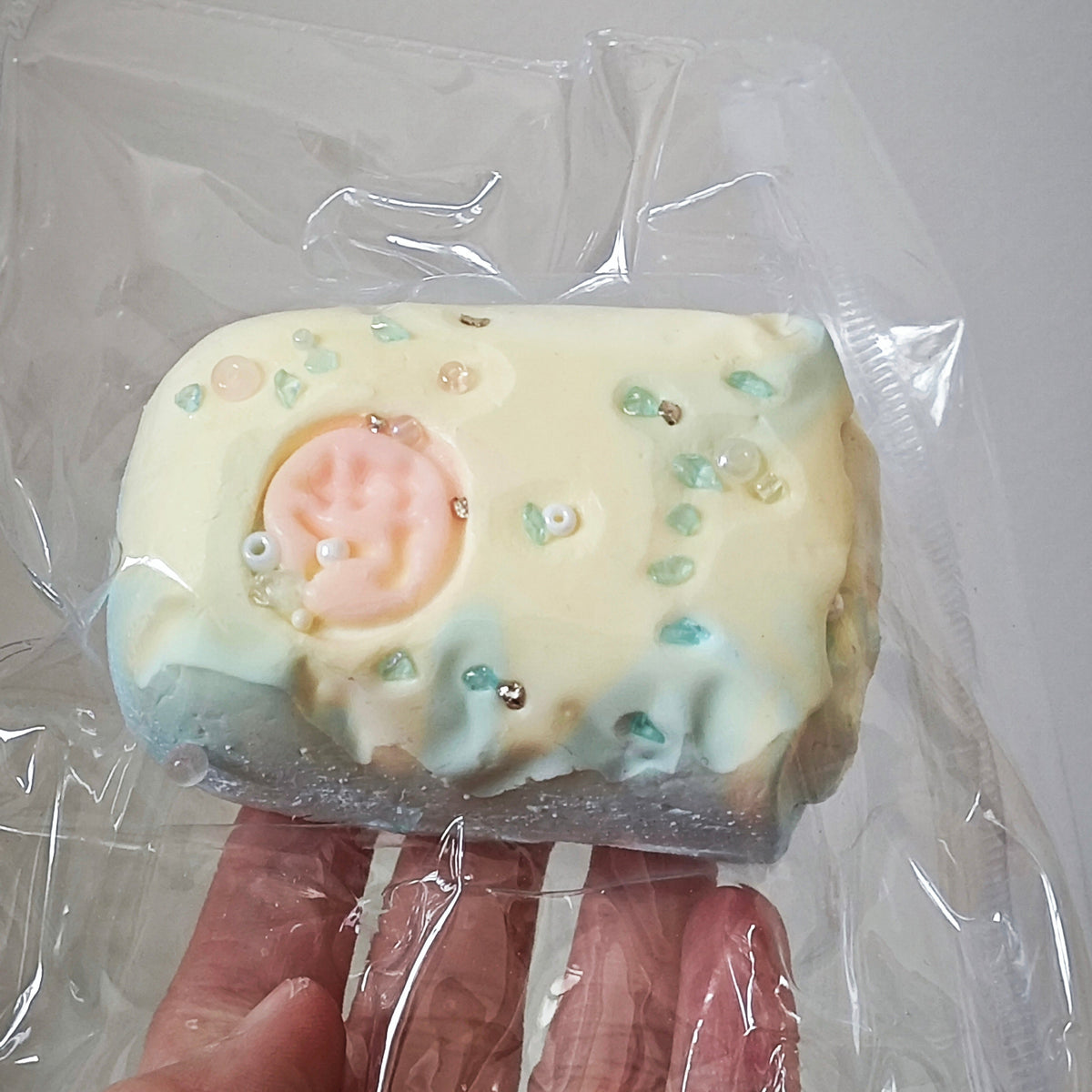 A hand holding a small rectangular pastel item wrapped in clear plastic. The item appears to be a decorative baked good or candy-like treat with pale yellow icing, mint green accents, tiny sprinkles, and a pink circular detail on top.