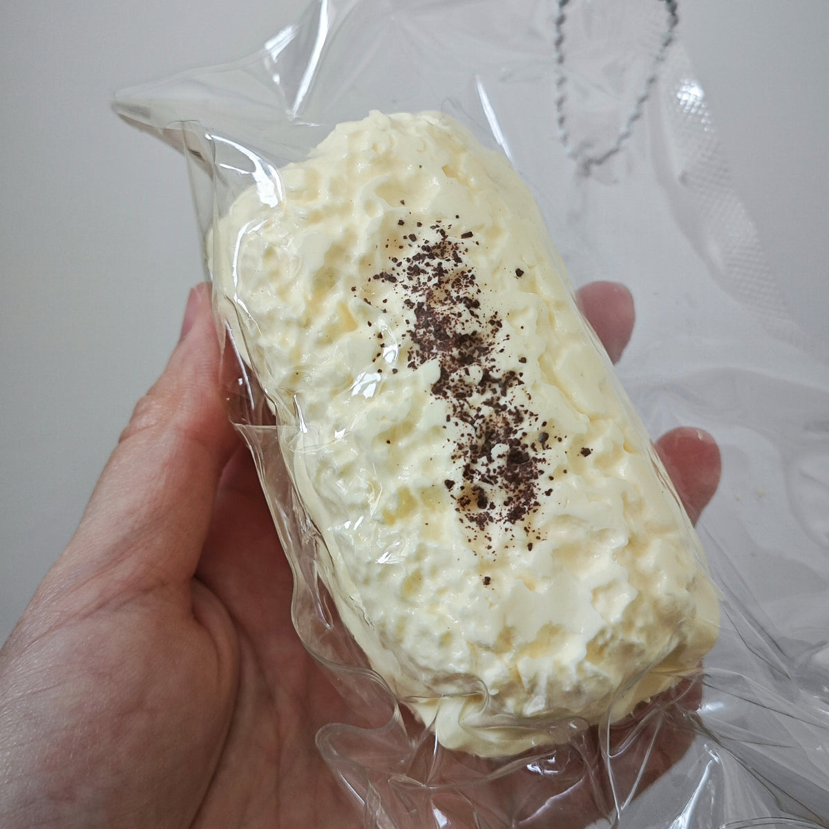 A hand holding a whipped cream-colored, loaf-shaped item wrapped in clear plastic, with a sprinkle of dark brown seasoning on top and a silver ball chain attached near the top right.