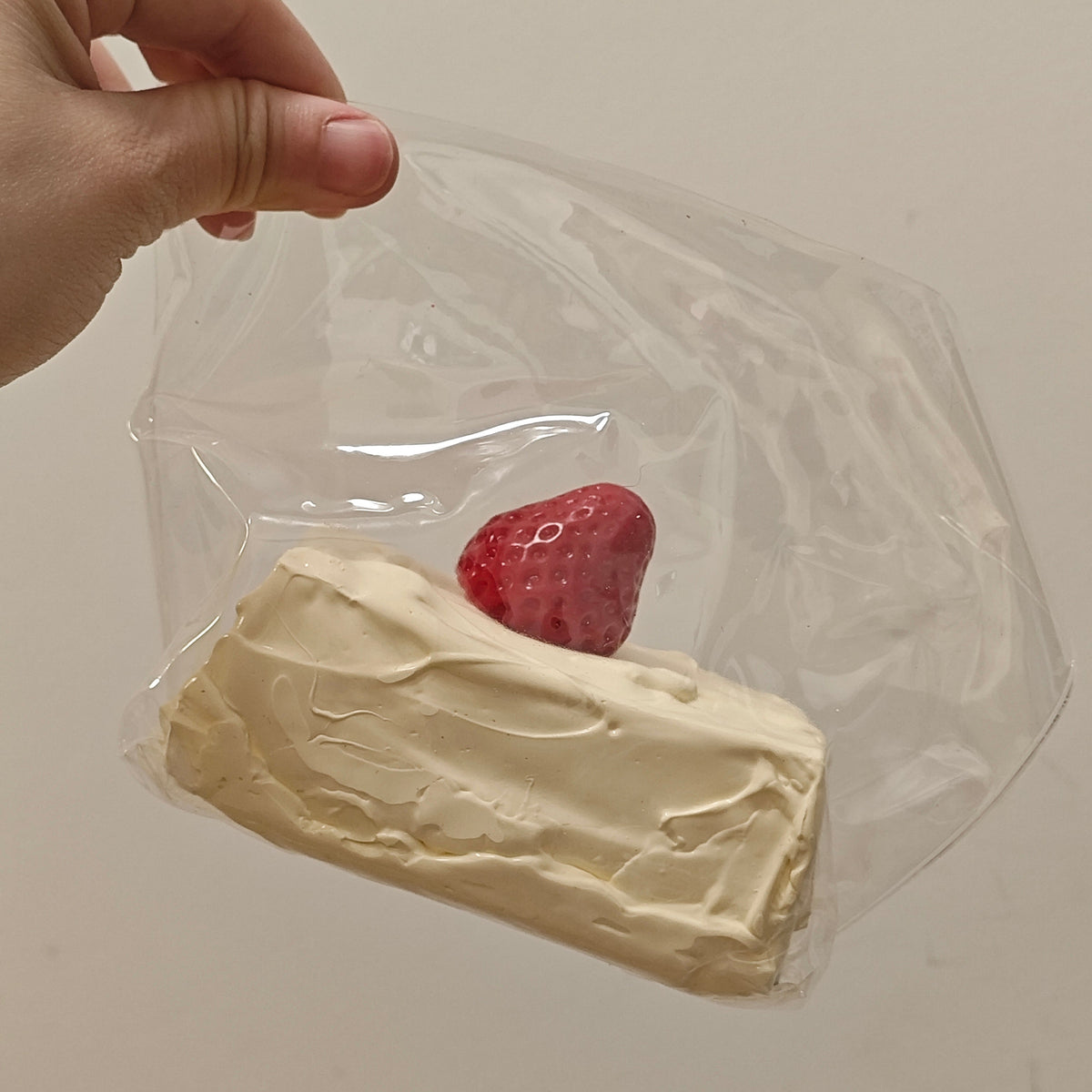 A hand holding a clear plastic bag containing a cream-colored cake slice with a red strawberry on top, set against a plain off-white background.
