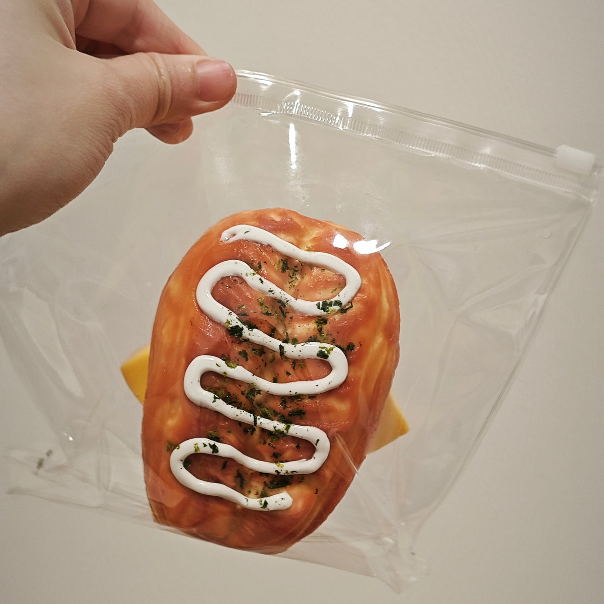A hand holds a clear plastic zip-top bag containing a novelty food item shaped like a sausage or hot dog with white drizzle and green herb garnish, against a plain light background.
