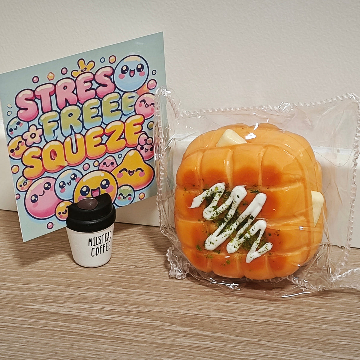An orange pumpkin-shaped squishy toy with white drizzle details, sealed in a clear plastic bag, displayed next to a 'Stress Free Squeeze' card and a miniature coffee cup on a wooden surface.