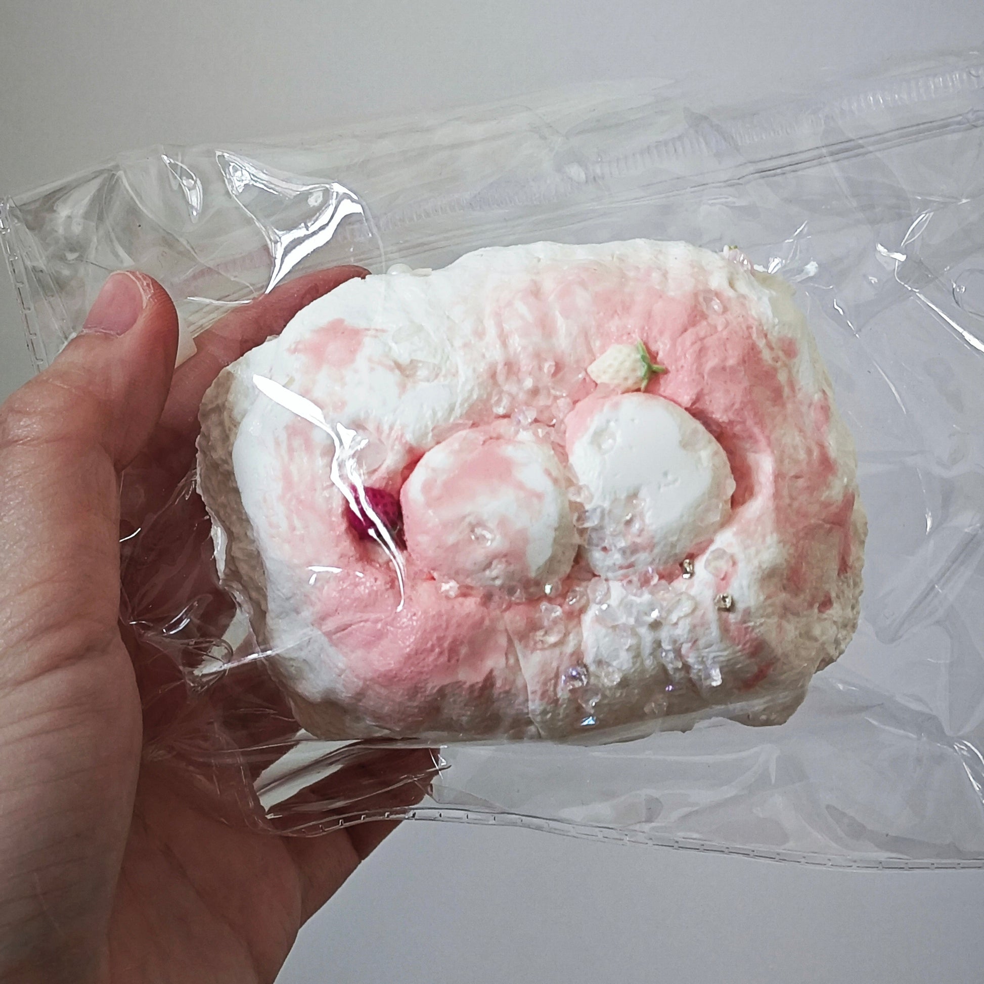 A hand holding a packaged squishy toy with a pink and white marshmallow-like design inside a clear plastic bag, against a plain light gray background.