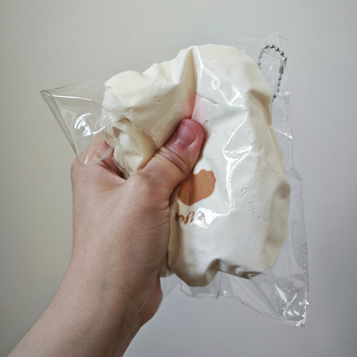 A hand holding a cream-colored fabric item inside a clear plastic bag against a plain light gray background.