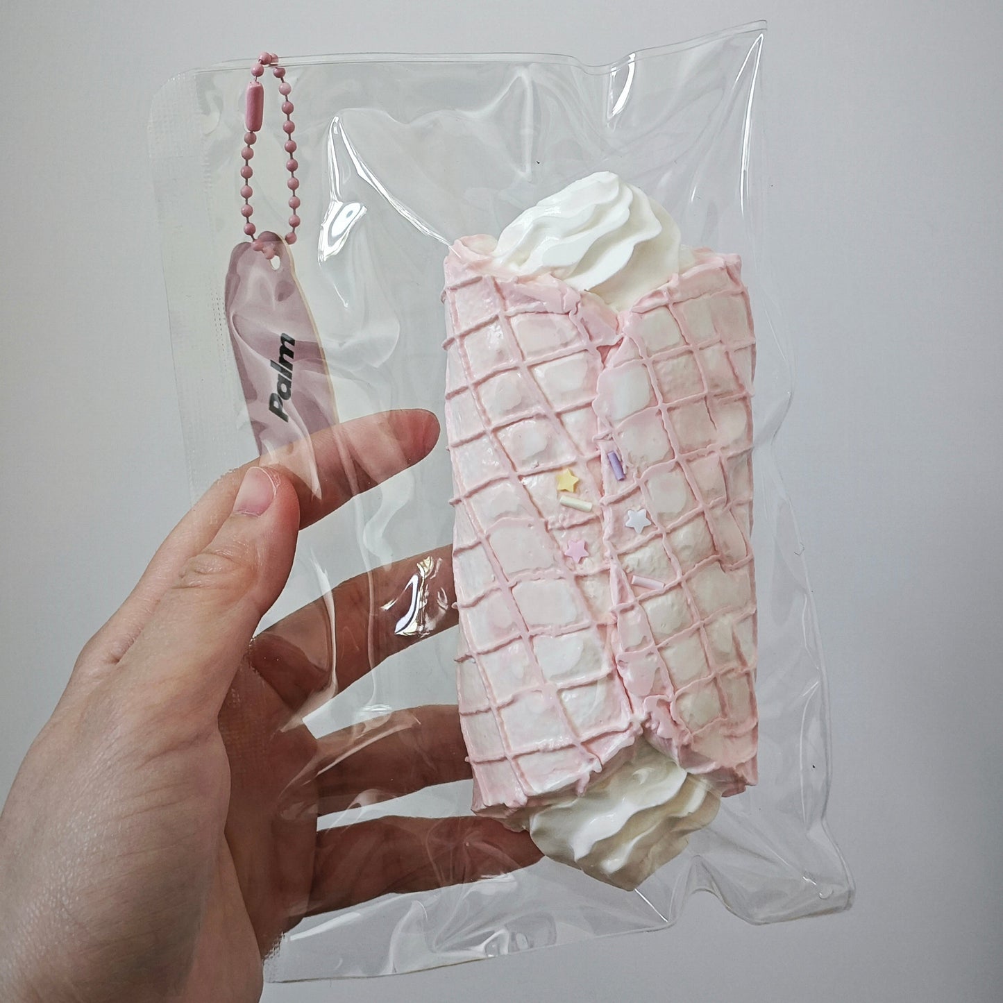 A hand holding a clear plastic bag containing a small novelty item shaped like a pink pastry or dessert with white cream tops. The bag has a pink ball chain keychain attached, and the background is a plain light gray wall.