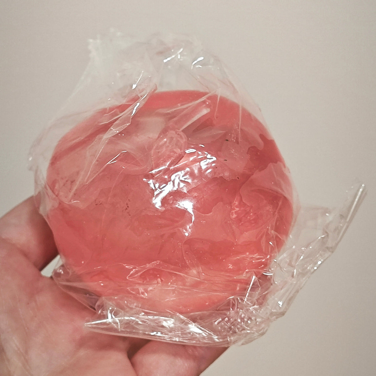 A hand holding a round, translucent pink item wrapped in clear plastic against a plain light beige background.