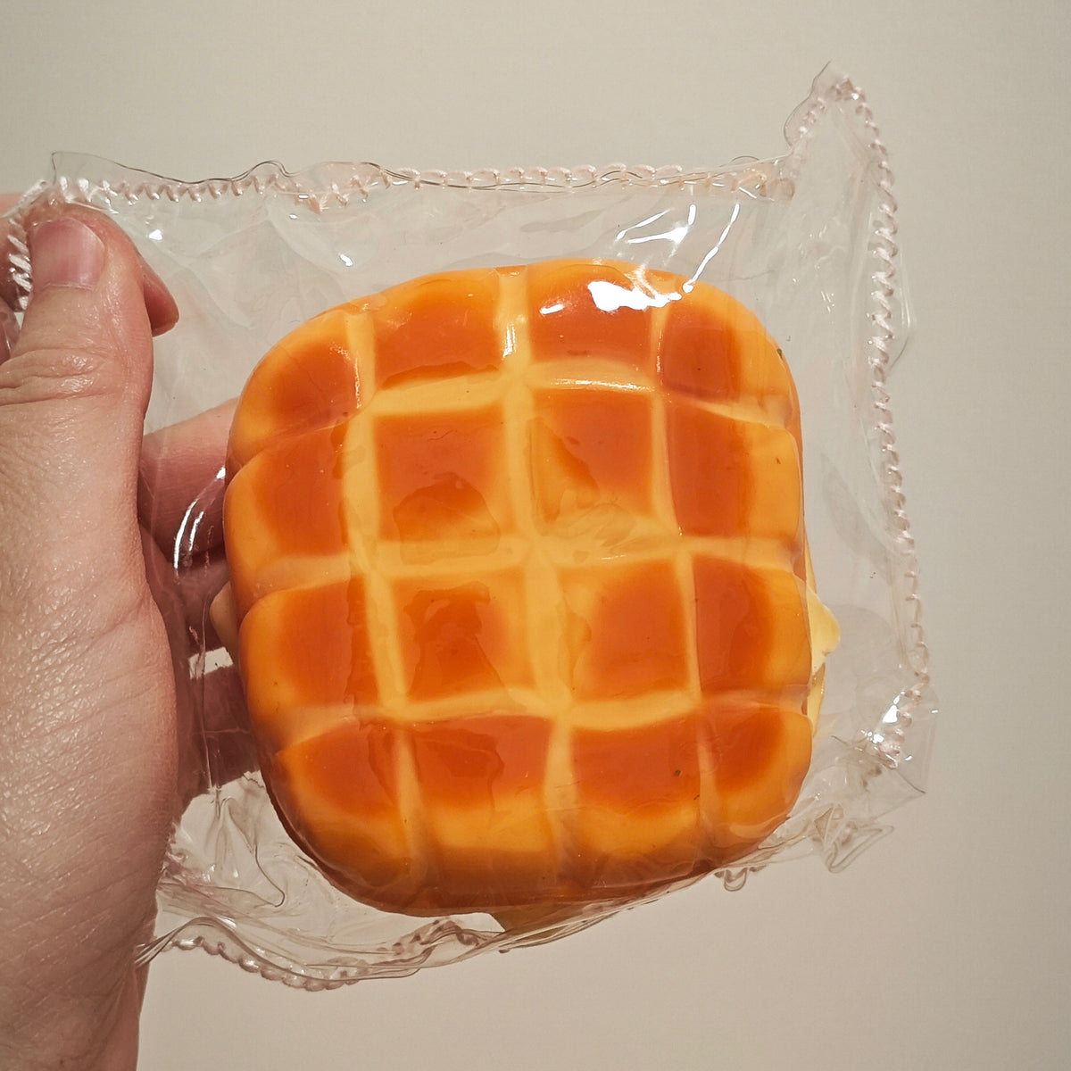 A hand holding a packaged orange-brown waffle-shaped squishy toy against a plain light beige background.