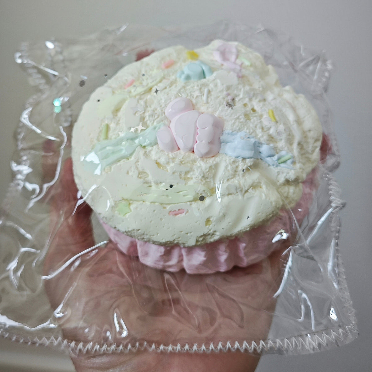 A hand holding a frosted cupcake-shaped item wrapped in clear plastic. The top is pale cream with pastel pink, blue, and green accents, and a pink ruffled base, against a plain light background.