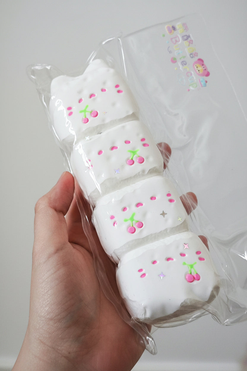 A hand holding a clear plastic bag containing four white rectangular marshmallows decorated with pink cherry and star details, against a plain light gray background.