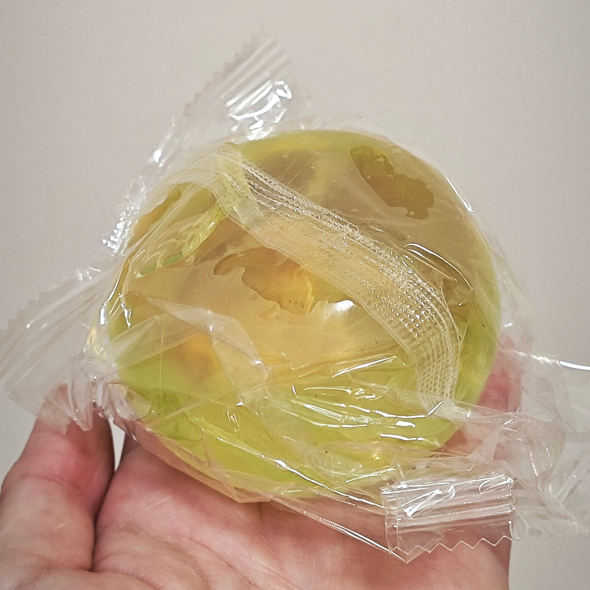 A hand holding a small, round, translucent yellow-green candy or gelatinous snack wrapped in clear plastic packaging against a plain light background.
