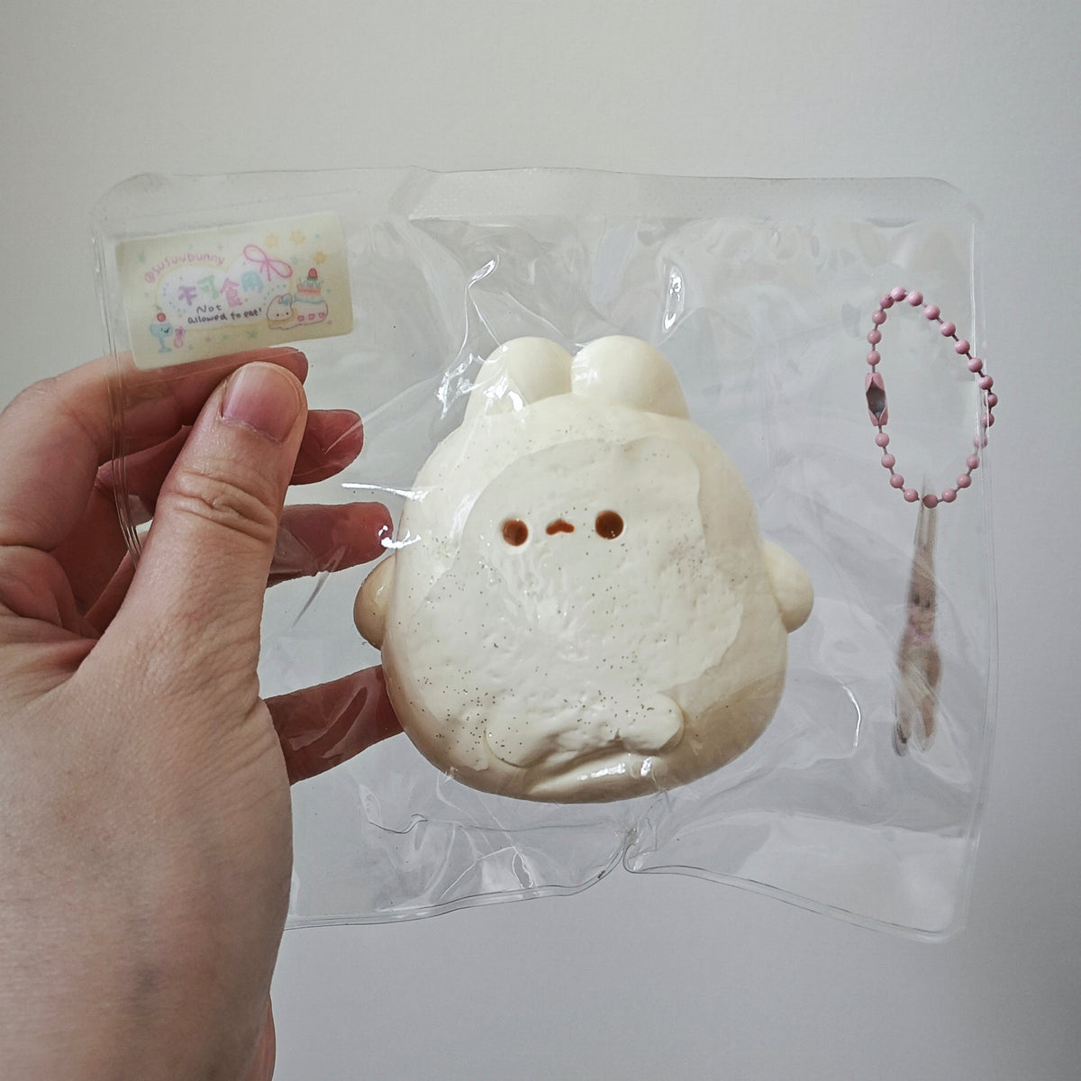 A hand holding a clear plastic bag containing a small, round, cream-colored plush toy with a simple face. The bag also contains a small tag and a pink beaded keychain.