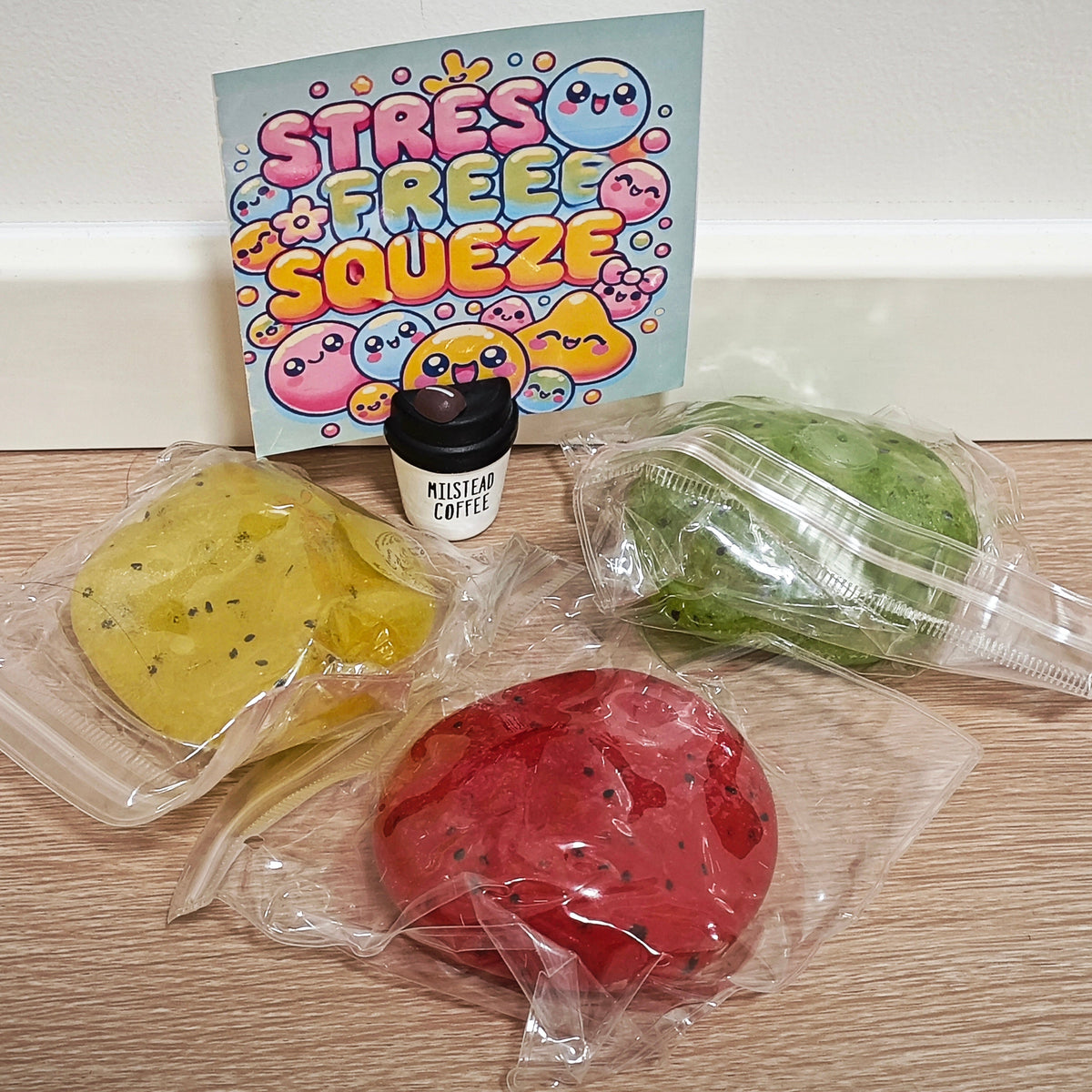 Three round squeeze toys in clear plastic bags on a wood surface, with a small black cup labeled Mission Coffee and a colorful card reading 'Stress Free Squeeze' behind them.