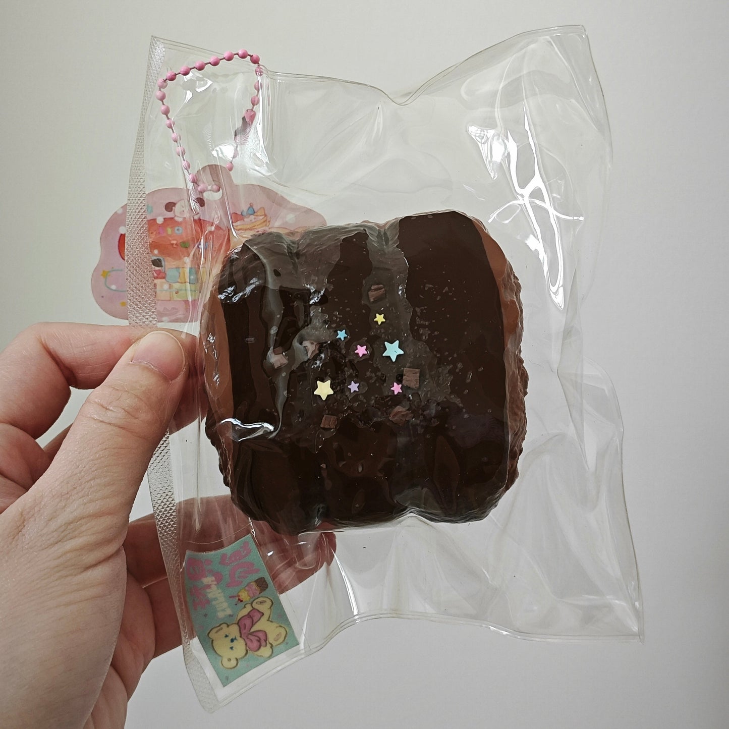 A hand holding a clear plastic bag containing a brown chocolate-shaped squishy toy with small multicolored star accents and a pink-and-white string charm, against a plain light background.