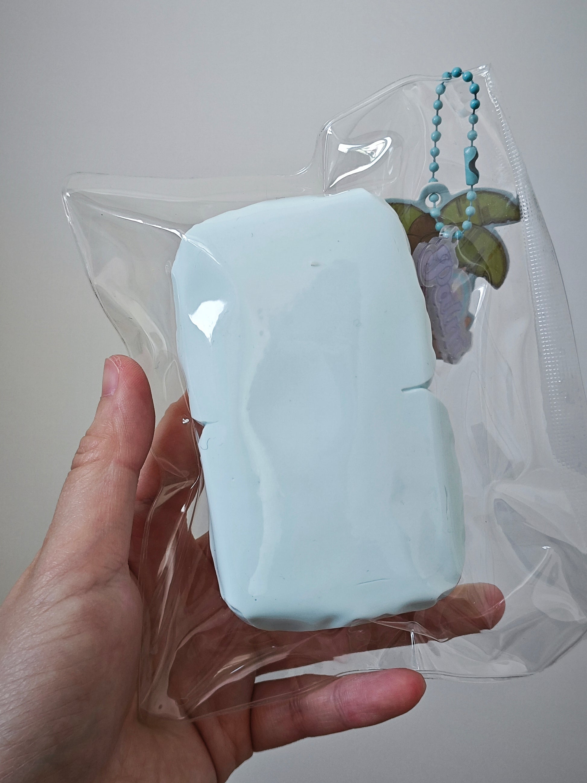 A hand holding a light blue rectangular item sealed in a clear plastic bag, with a small hanging charm attached near the top right.