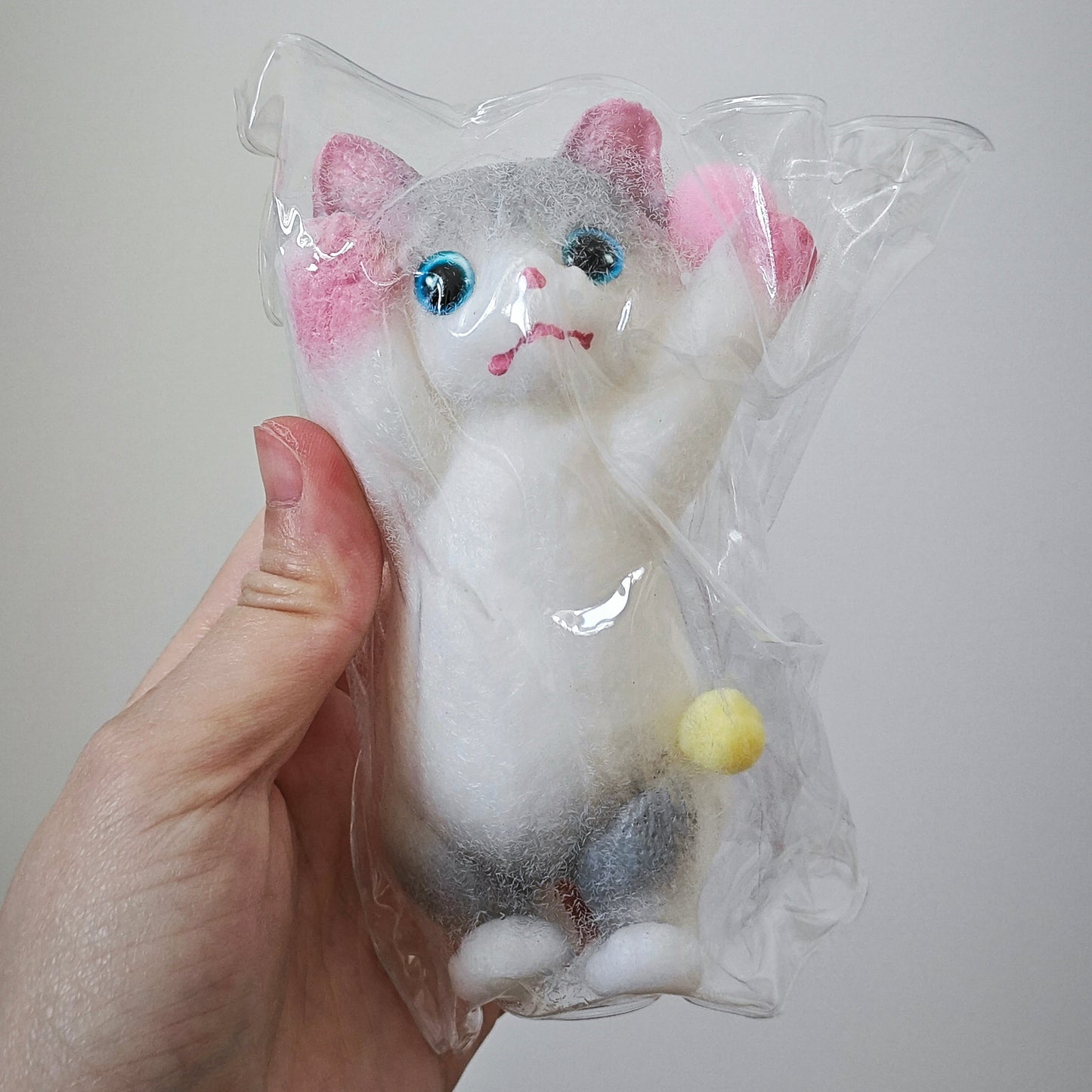 A hand holding a small felted animal toy shaped like a cat inside a clear plastic bag. The toy is white with gray patches, pink inner ears, blue eyes, and a small yellow tail, photographed against a plain light gray background.