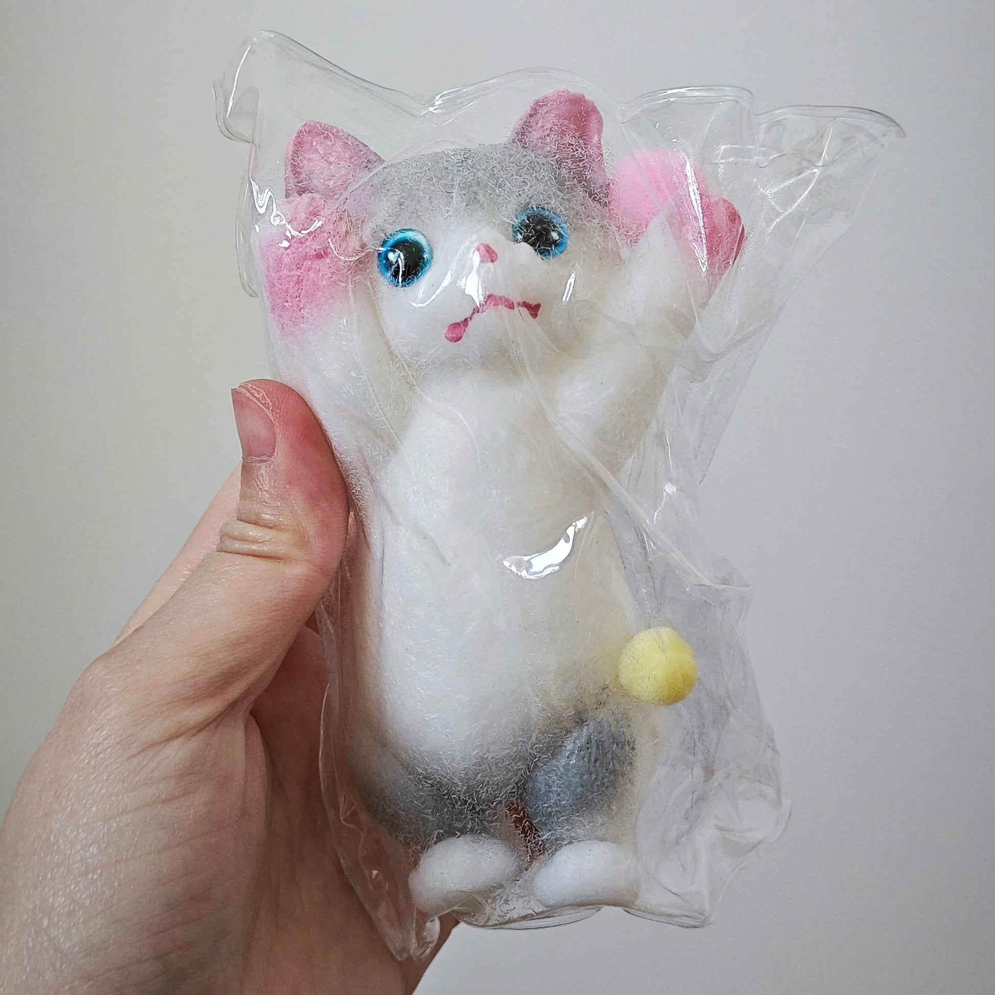 A hand holding a small felted animal toy shaped like a cat inside a clear plastic bag. The toy is white with gray patches, pink inner ears, blue eyes, and a small yellow tail, photographed against a plain light gray background.