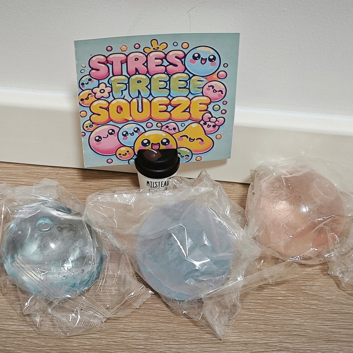 Three pastel-colored stress balls in clear plastic packaging on a wooden floor, with a colorful 'Stress Free Squeeze' card behind them.