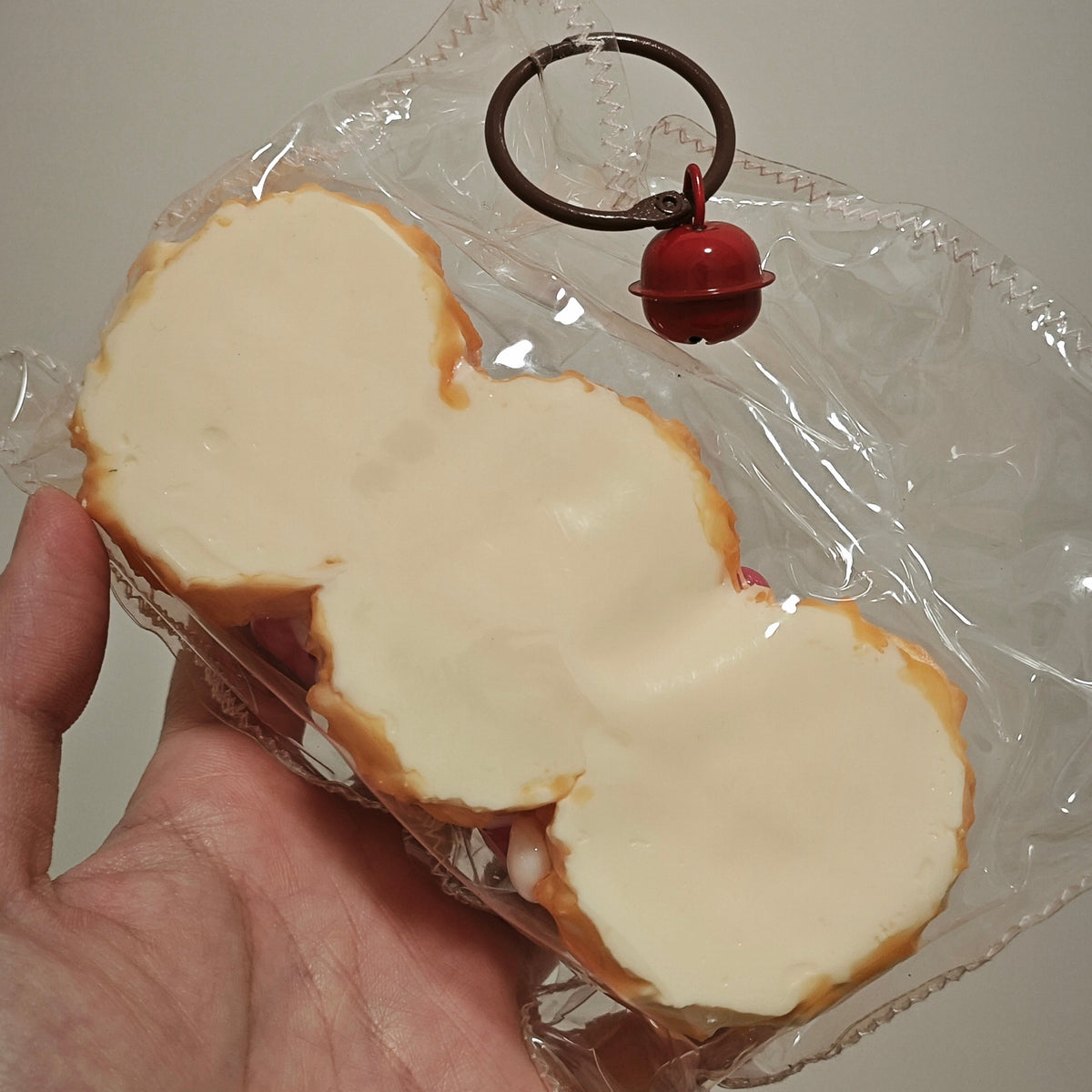 A hand holding a packaged novelty item shaped like a snowman, featuring a cream-colored center, tan edges, and a small red bell attached to a black ring through clear plastic wrapping.